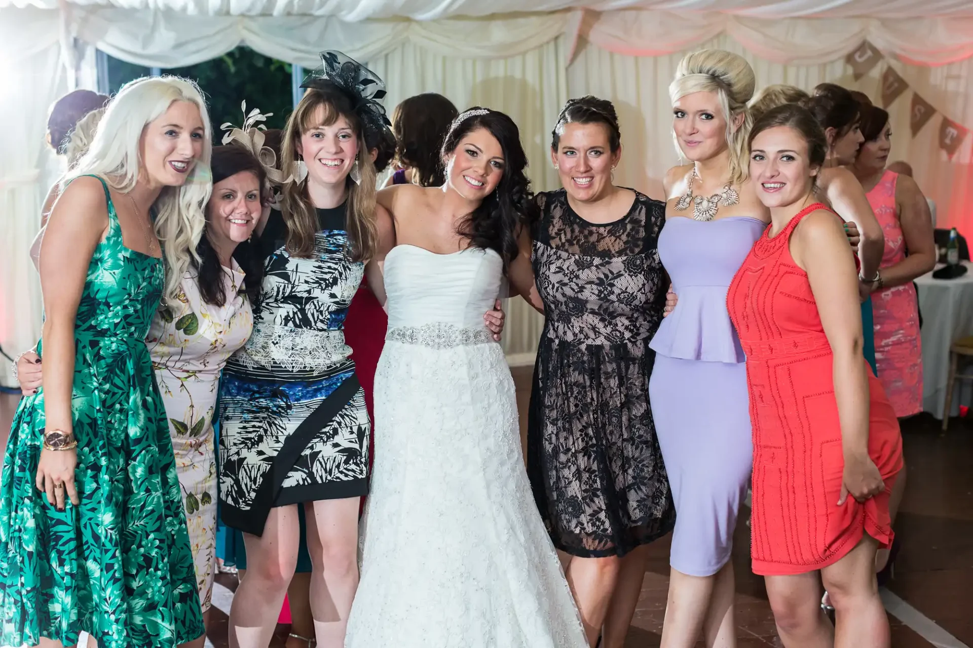 A bride in a white dress surrounded by six women in various colorful dresses, smiling together at a wedding reception.