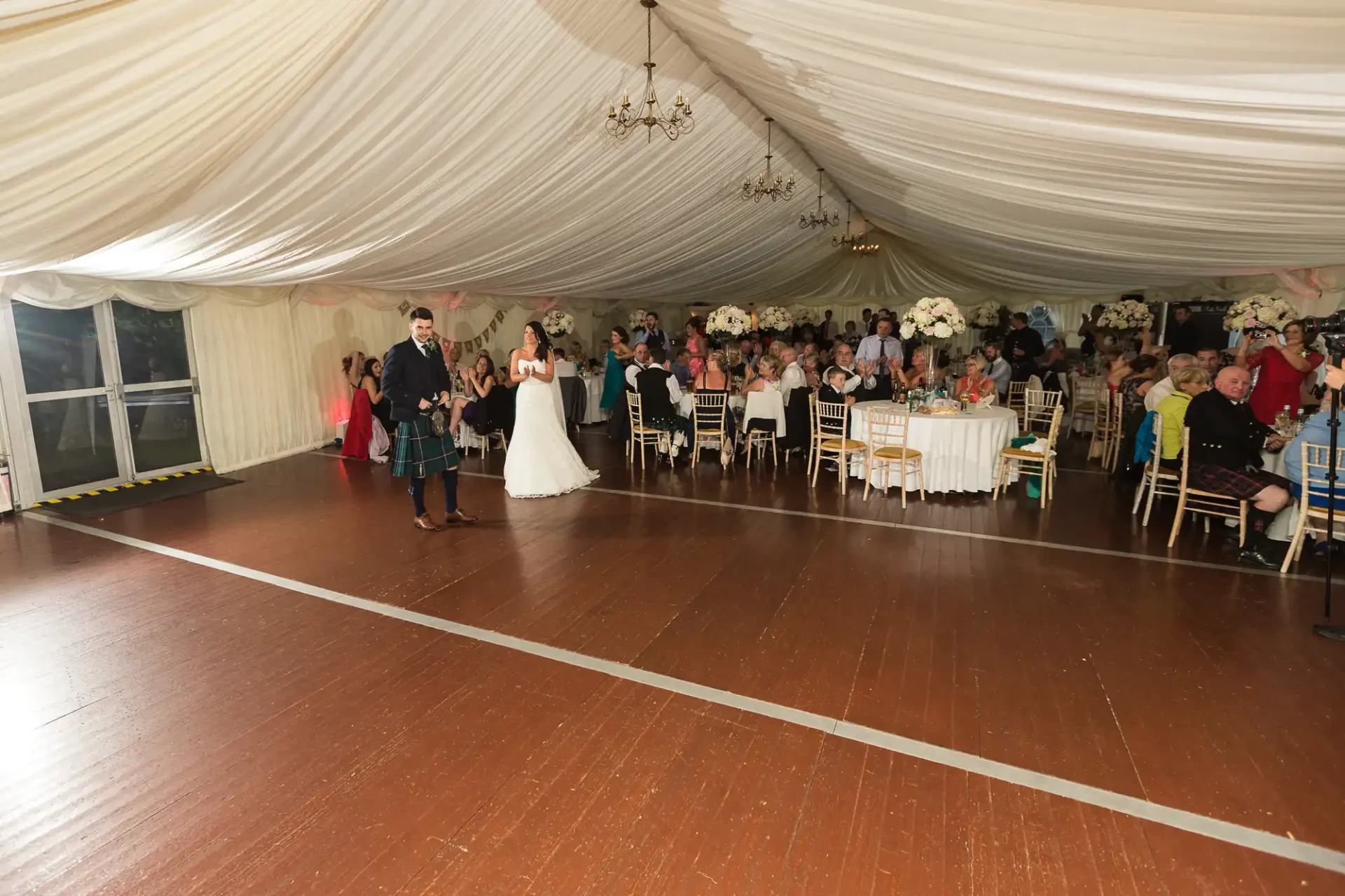 A bride and groom walking through a tent reception area with guests seated around, clapping and celebrating.