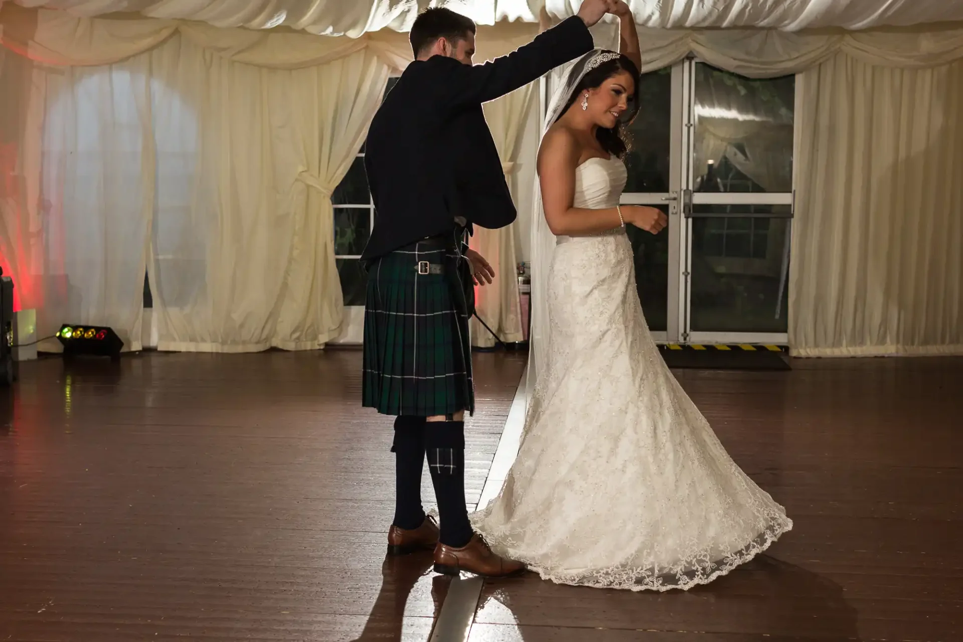 A bride and groom share a dance at their wedding reception, the groom wearing a traditional kilt.