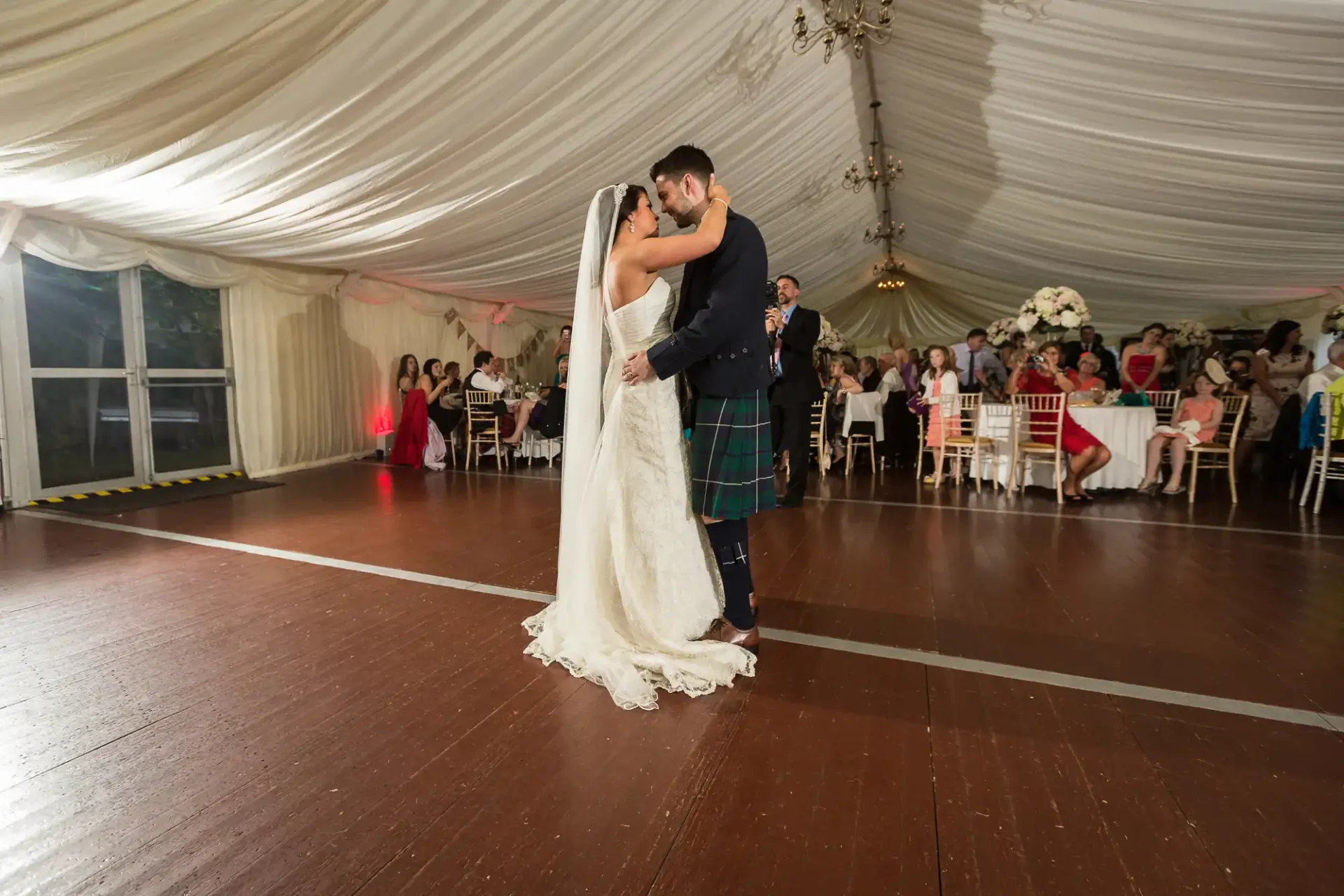 A bride and groom share a dance wearing wedding attire; the groom is in a kilt. guests seated around them in a tented venue.