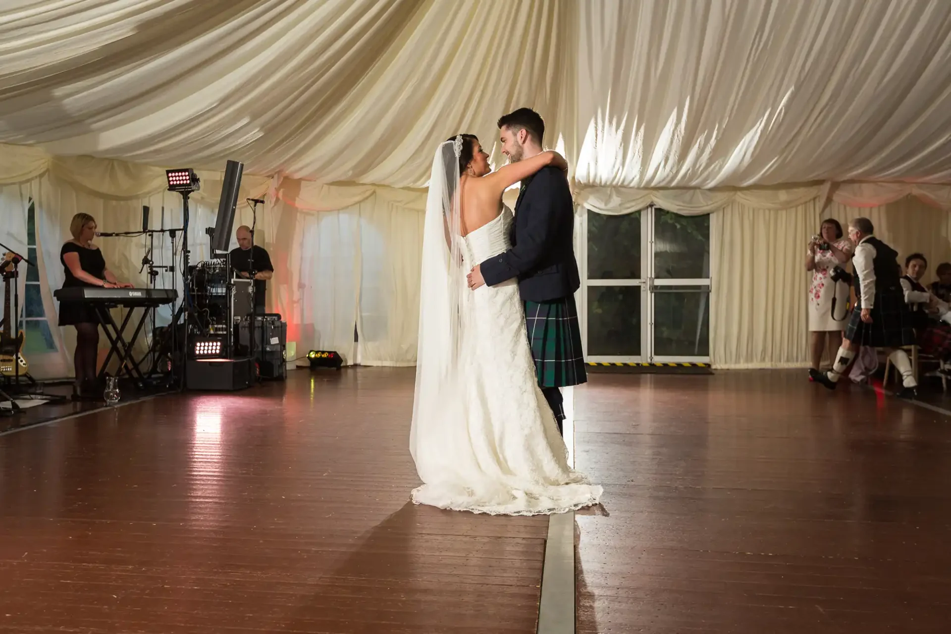 A bride in a white gown and a groom in a kilt embrace while dancing in a marquee, with a live band and guests in the background.