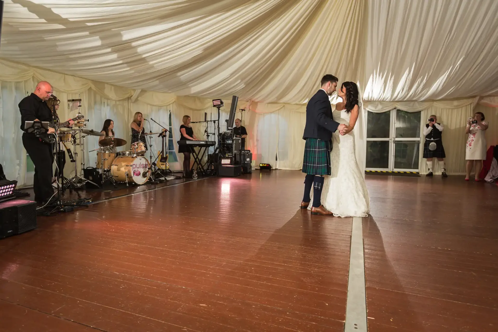 A bride and groom share a dance in a tented venue while a band performs in the background and guests watch.