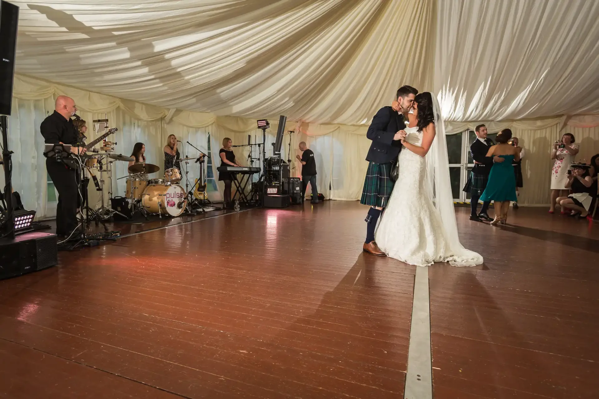 A bride and groom share a dance in a tent with a live band playing in the background and guests dancing around them.