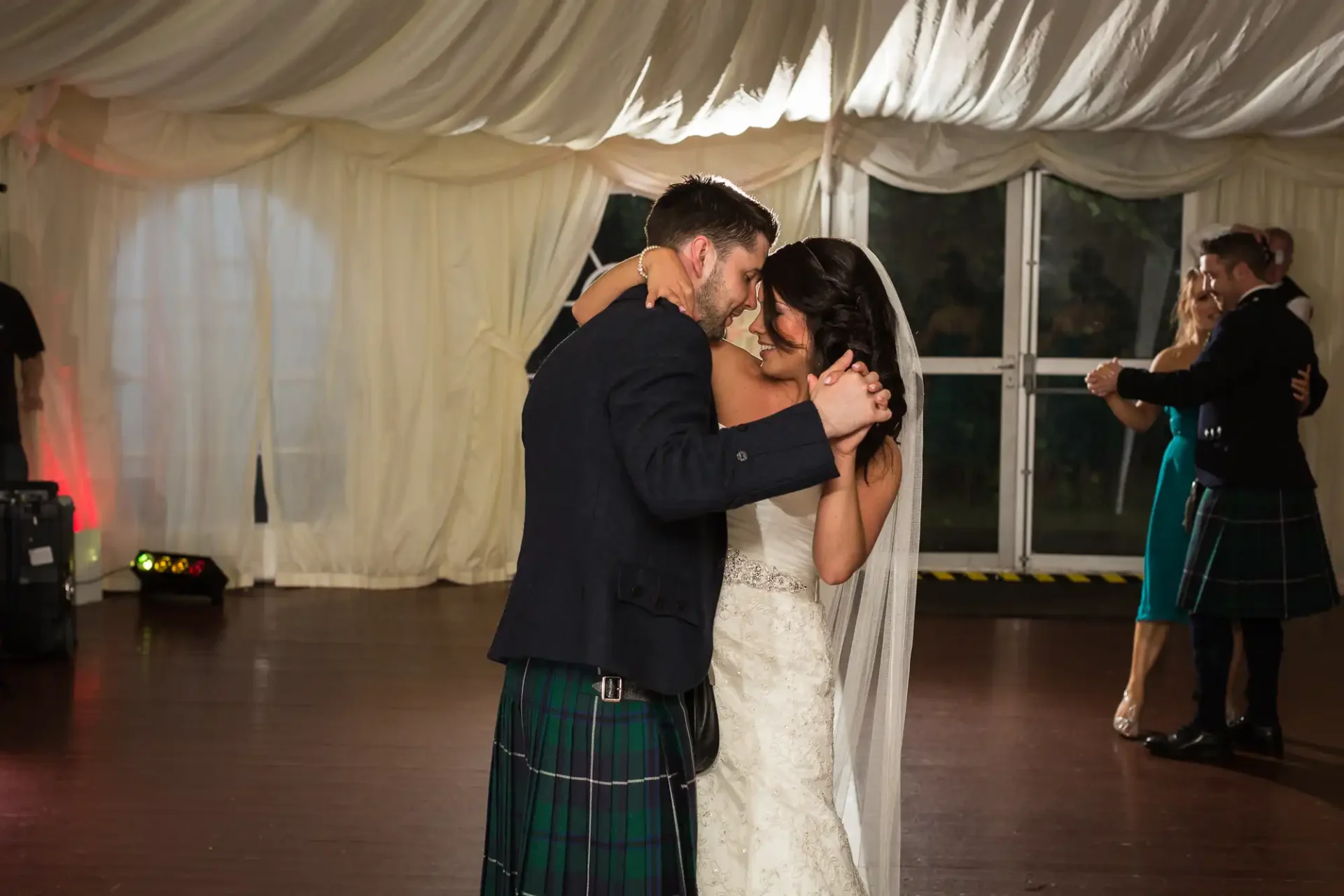 Bride and groom share a close dance at their wedding reception, with guests dancing in the background under a tented venue. the groom is wearing a kilt.