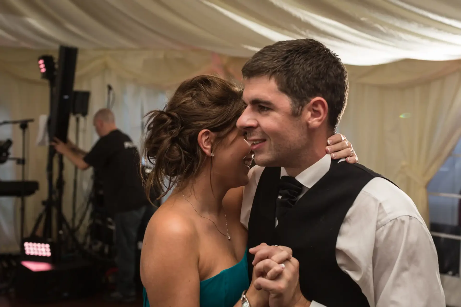 A couple dancing closely at a wedding reception with a dj performing in the background under a tent.