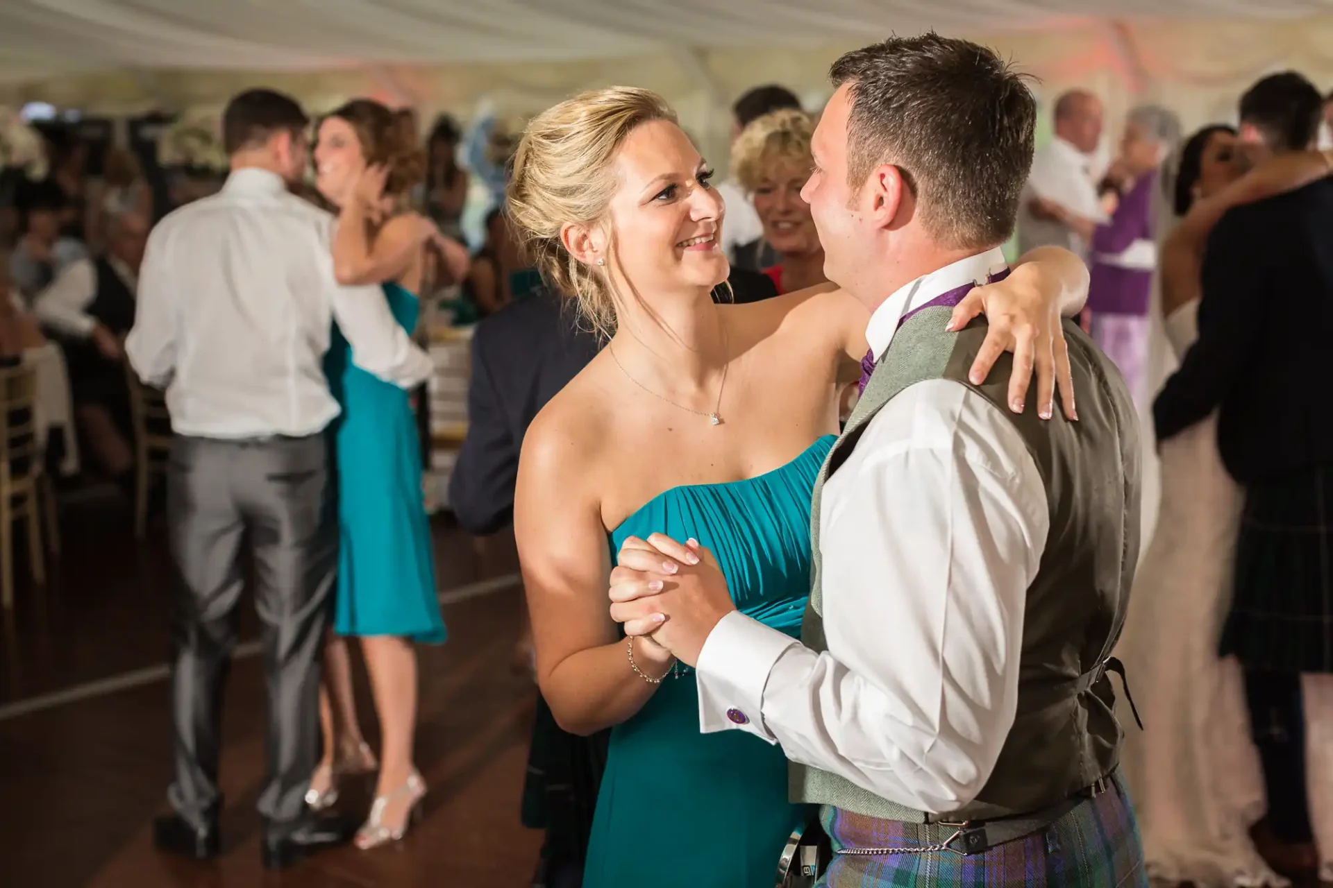 A couple dances closely, smiling at each other, at a wedding reception inside a tent filled with dancing guests.