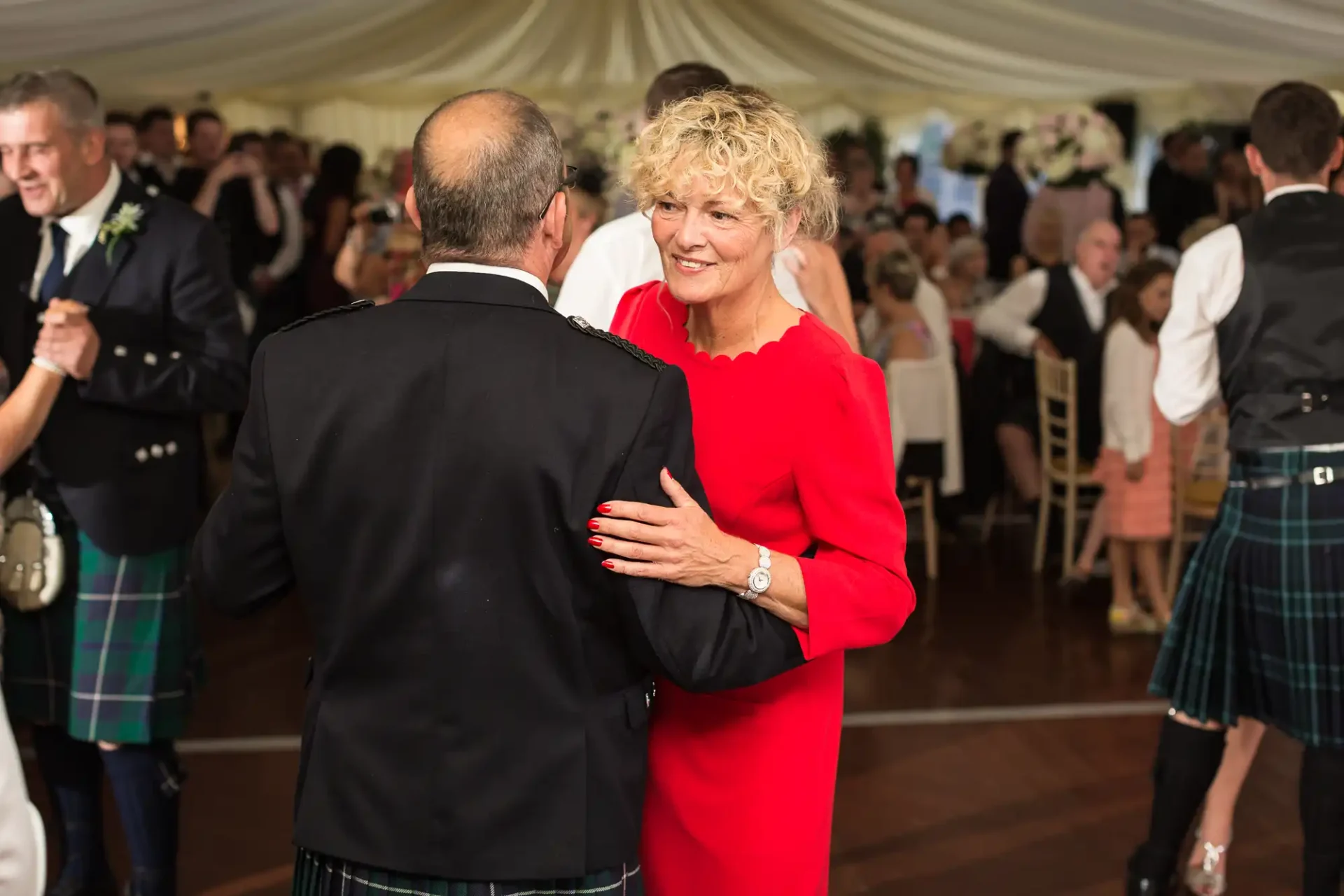 A woman in a red dress dancing with a man in a kilt at a lively wedding reception.