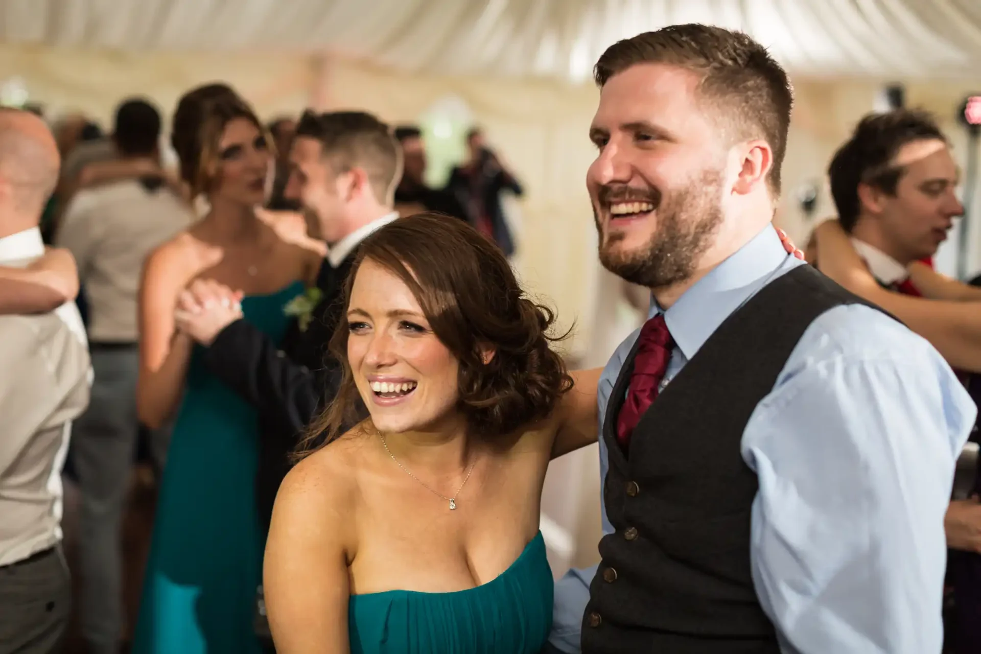 A joyful woman in a green dress and a man in a gray vest and red tie laughing together at a lively wedding reception.