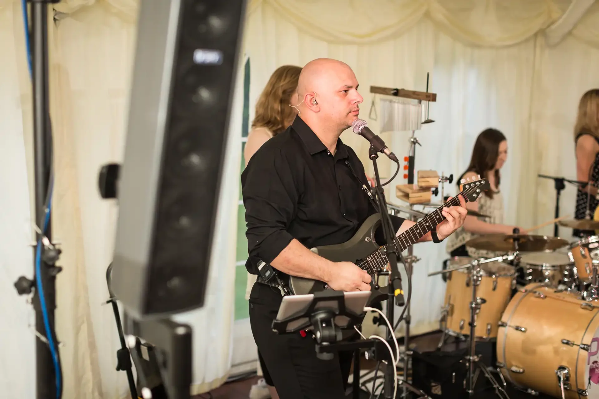 Bald musician playing electric guitar on stage at an event, with a drummer in the background.