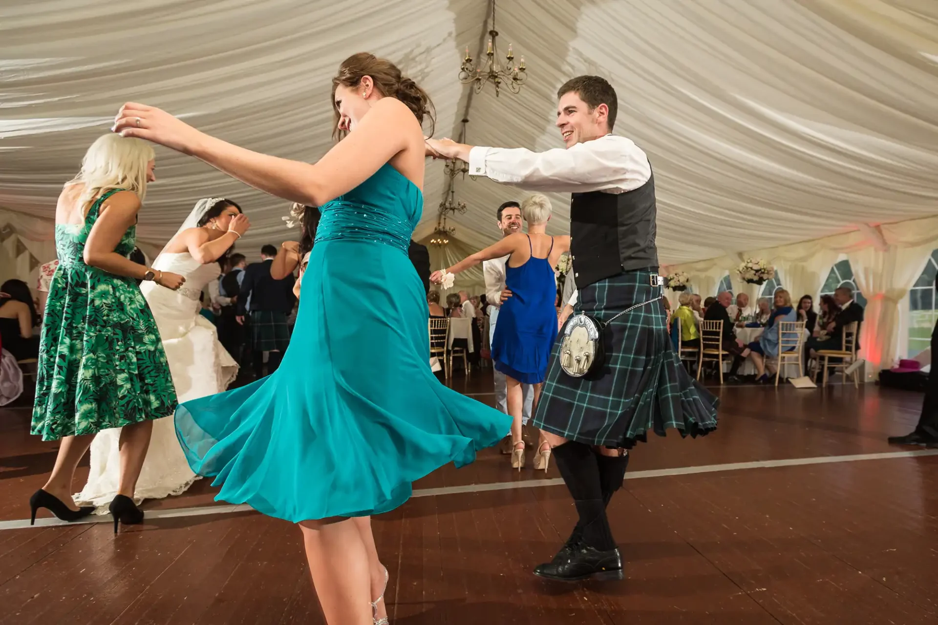 A couple dancing in a marquee, the woman in a teal dress and the man in a kilt, with guests seated in the background.