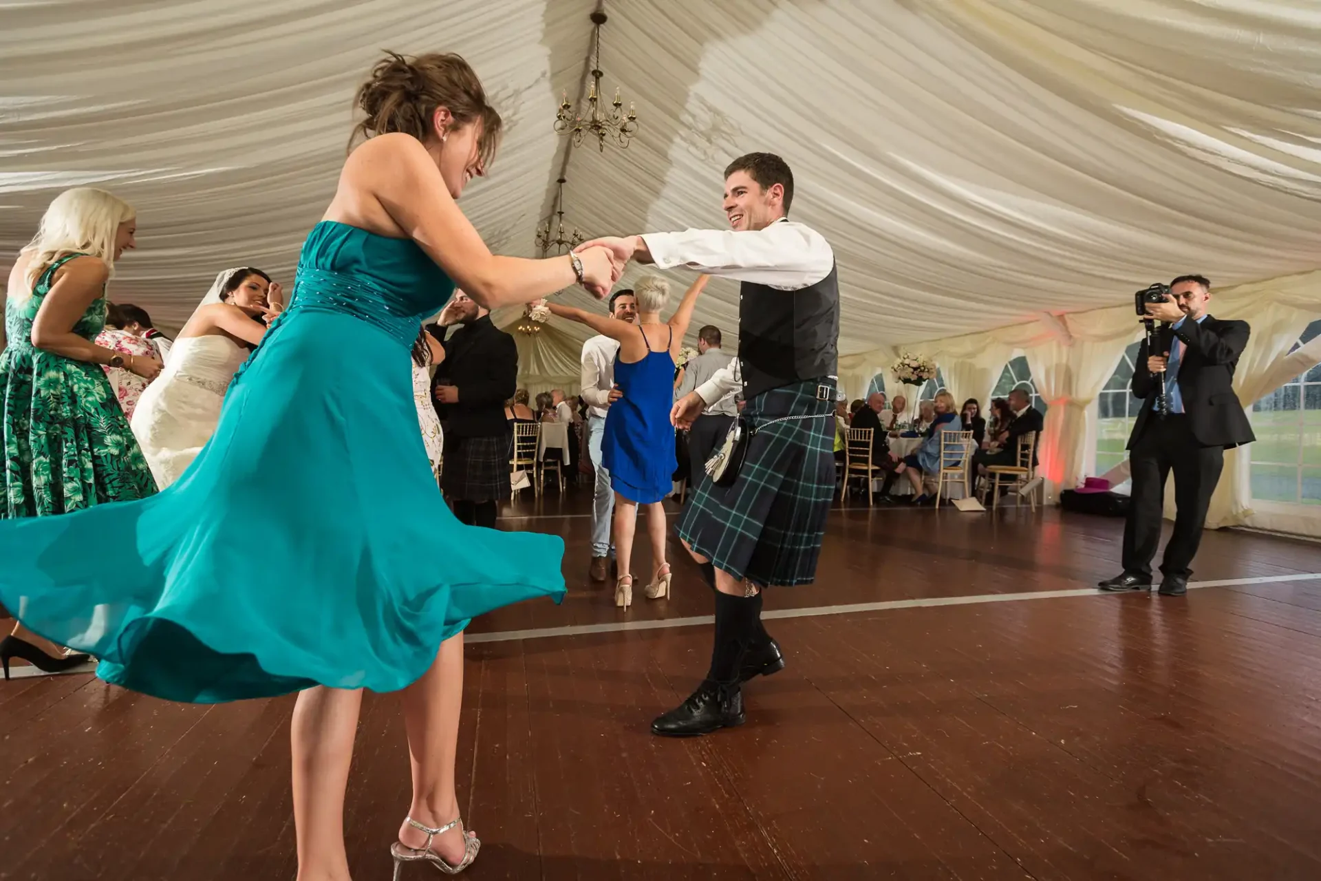 A couple dances joyfully in a tent at a wedding reception, the man in a kilt and the woman in a teal dress, while guests and a photographer watch.