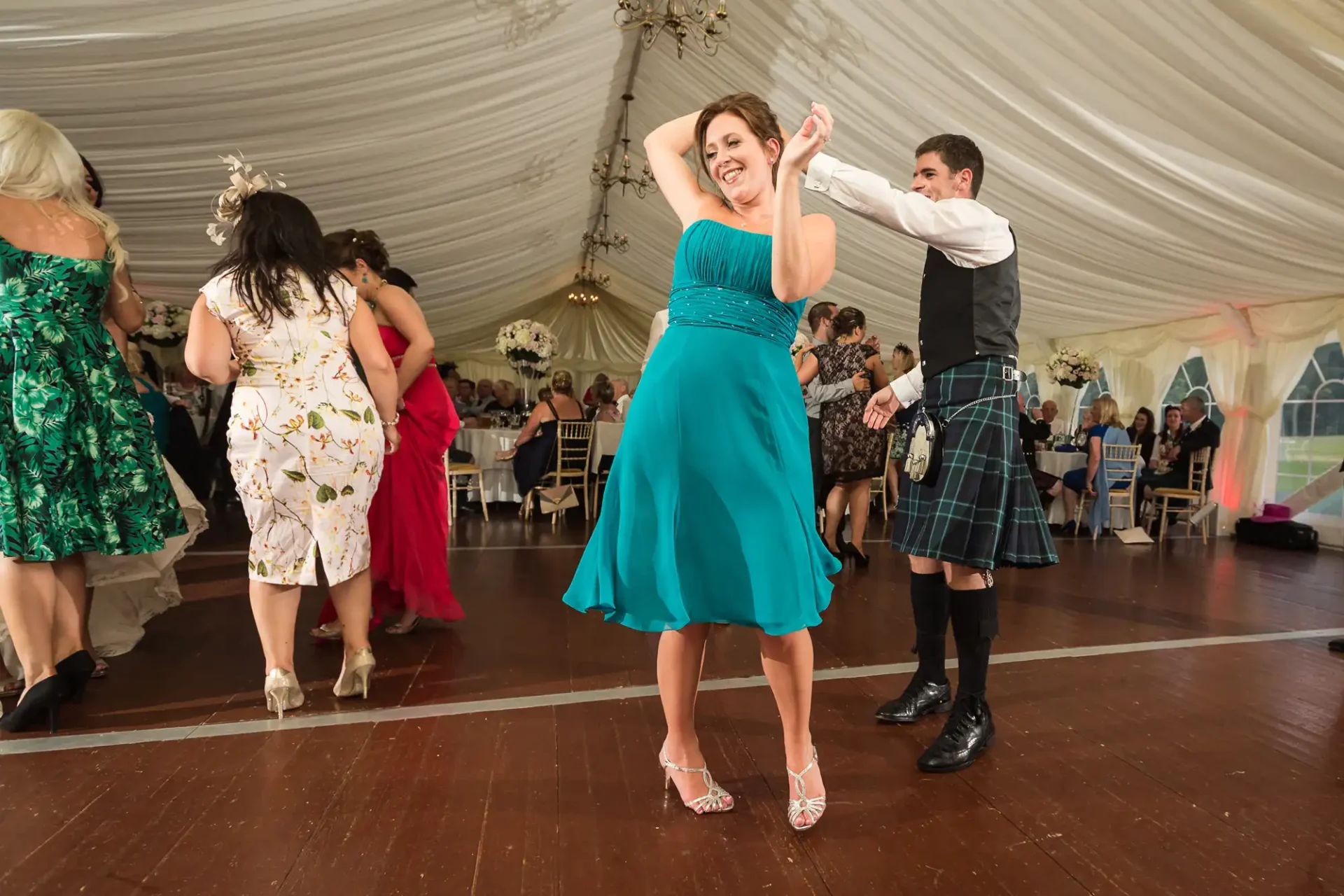 A woman in a turquoise dress dances joyfully with raised arms, and a man in a kilt dances behind her in a tent at a lively event.