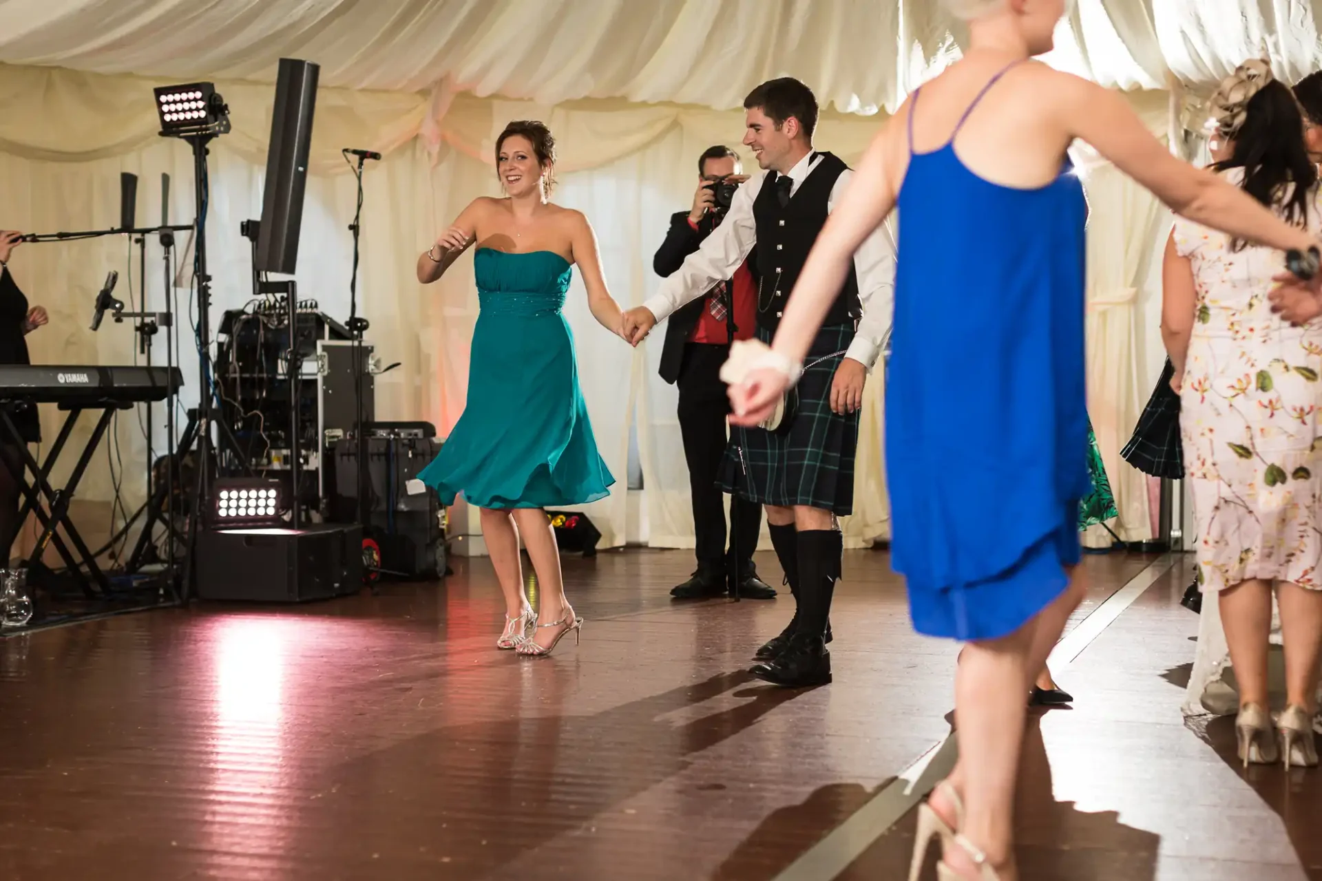 A woman in a teal dress dances with a man in a kilt at a lively wedding reception inside a tent.