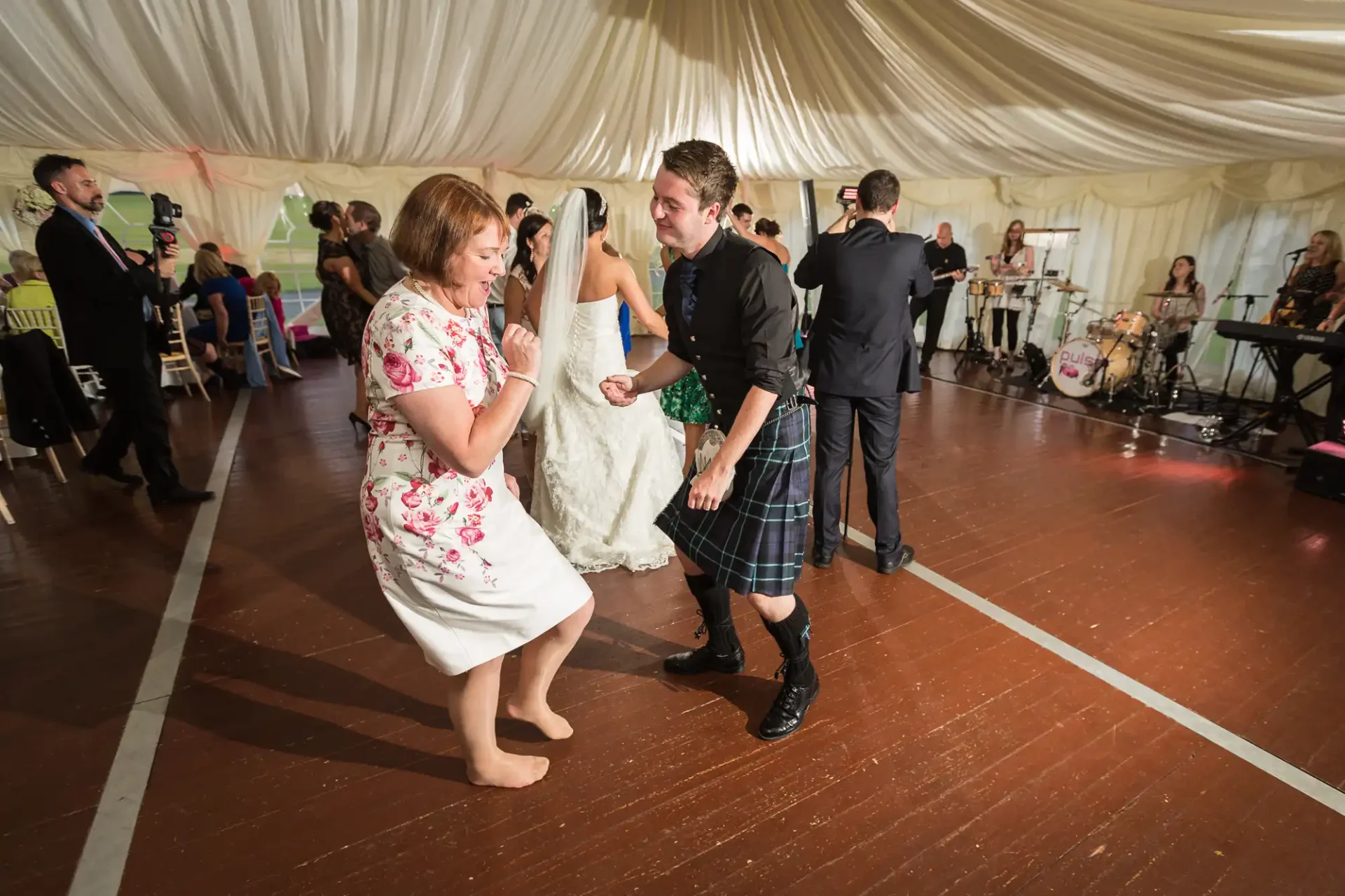 A man in a kilt dances with a woman in a floral dress at a lively wedding reception inside a marquee, with guests and a live band in the background.