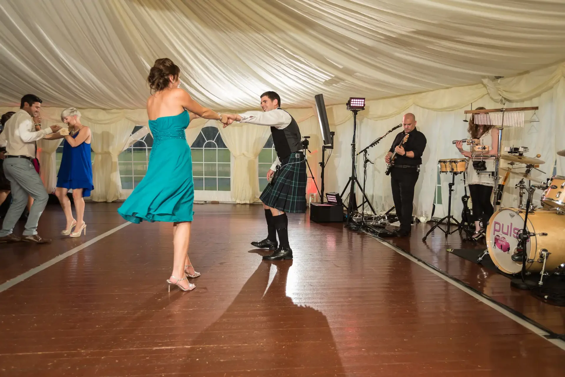 A couple dances energetically in a tent at a wedding, with the man in a kilt and the woman in a teal dress, while a band performs in the background.