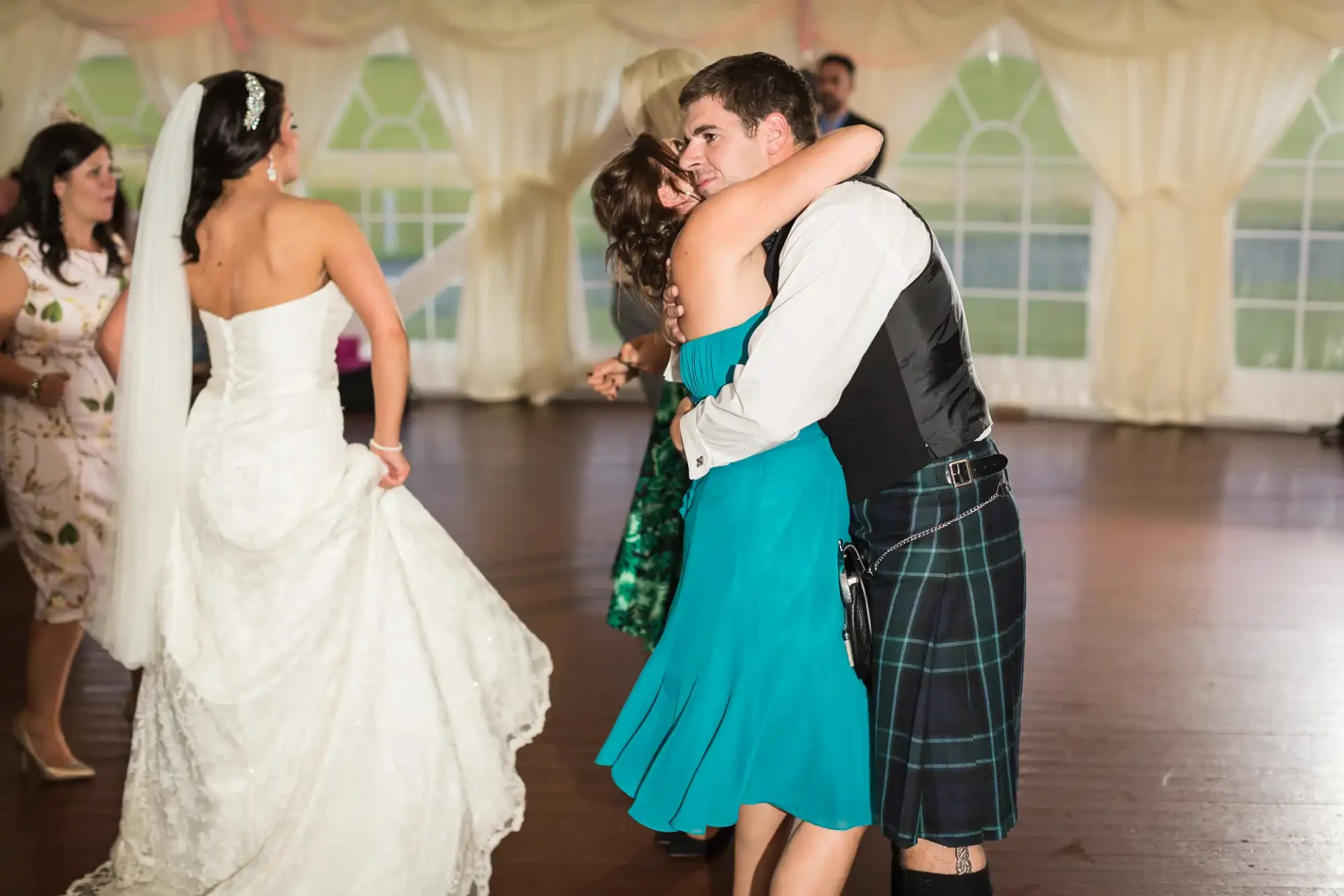 A bride in a white dress and a man in a kilt embracing on the dance floor at a wedding reception, with guests in the background.