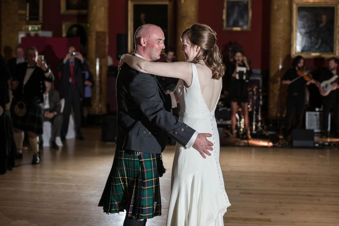 A couple dances closely in a grand hall, the man in a tartan kilt and the woman in a white dress, with a live band playing in the background.