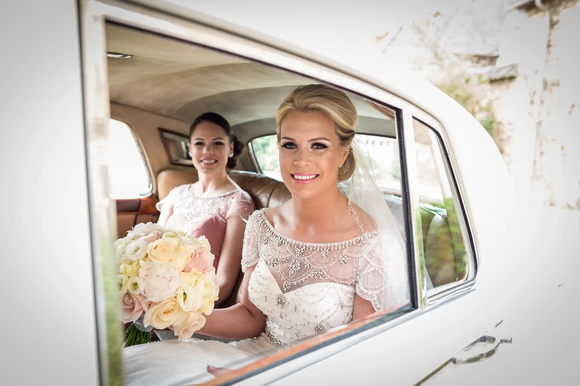 A bride holding a bouquet of flowers and her bridesmaid sit inside a car. The bride is smiling at the camera.