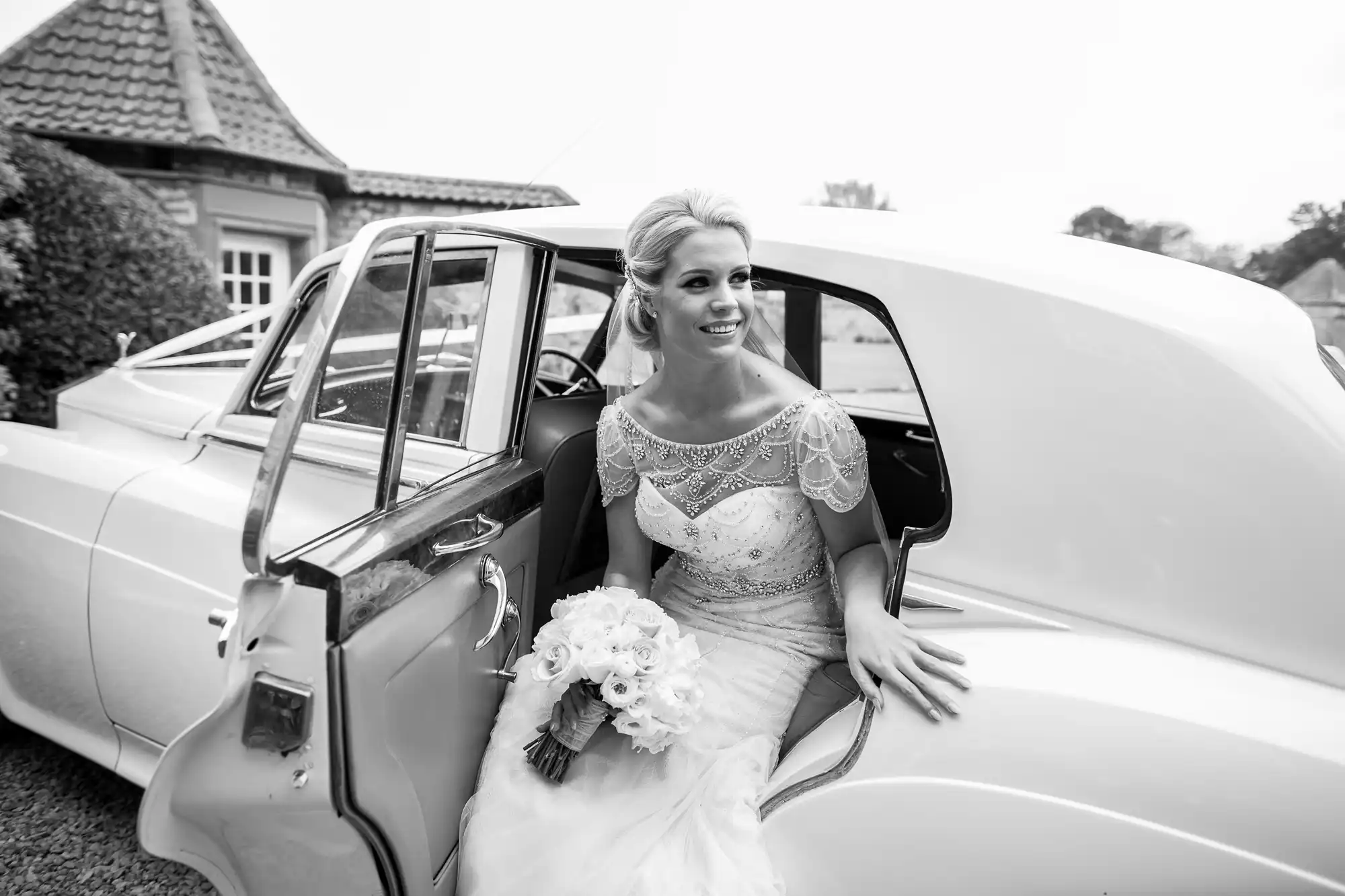 A bride wearing a lace wedding gown and holding a bouquet of flowers steps out of a vintage car on her wedding day.