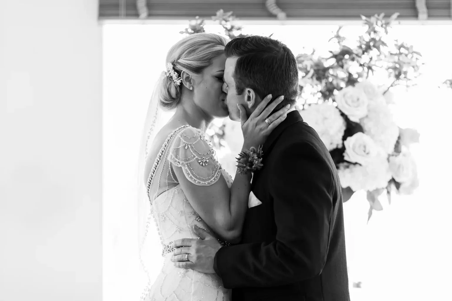 A newlywed couple dressed in wedding attire shares a kiss in front of a floral arrangement. The bride has her hair styled up with a hairpiece, and the groom is in a dark suit.