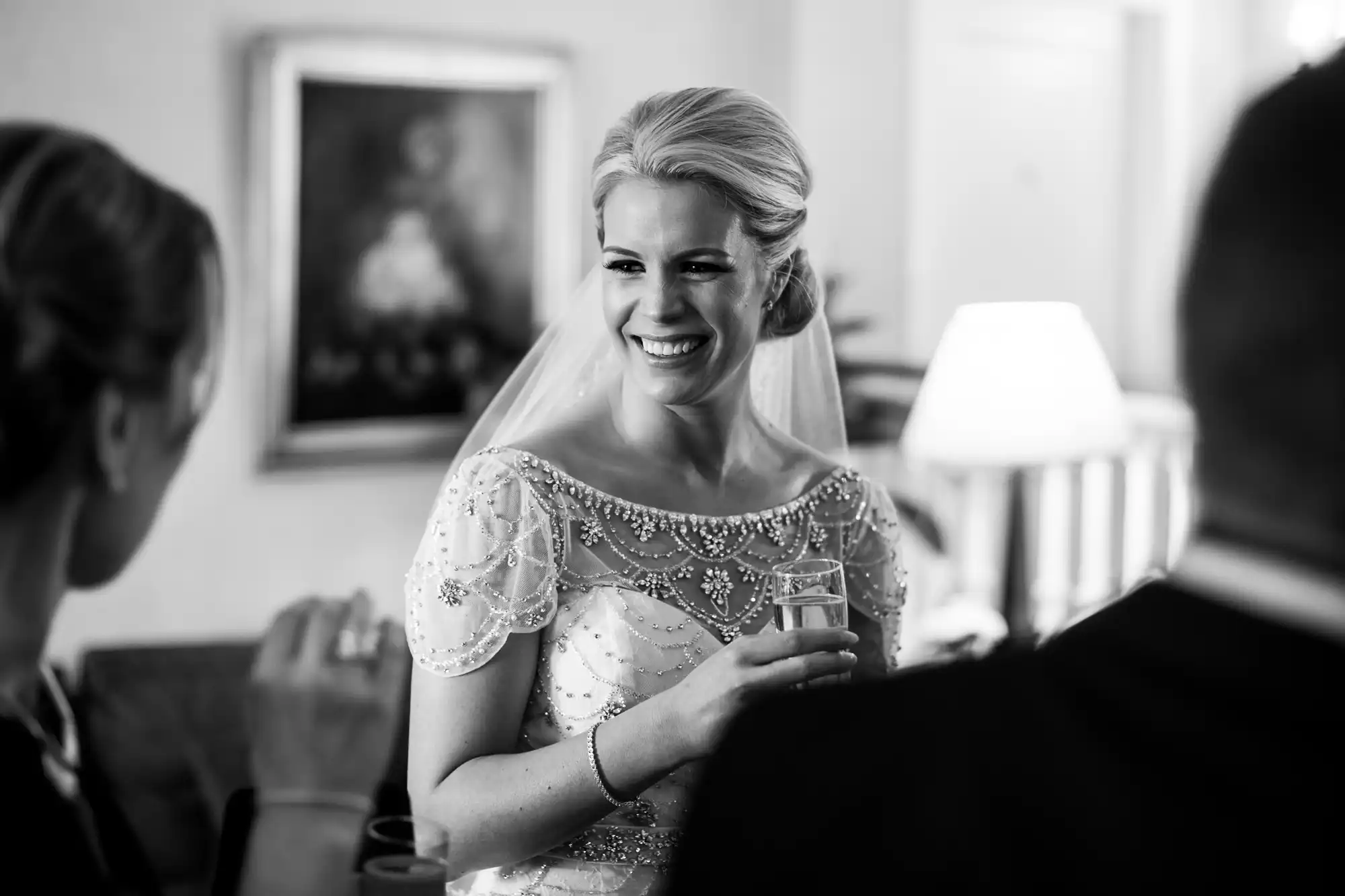 A smiling bride in a beaded dress holds a drink while conversing with guests in a well-lit room.