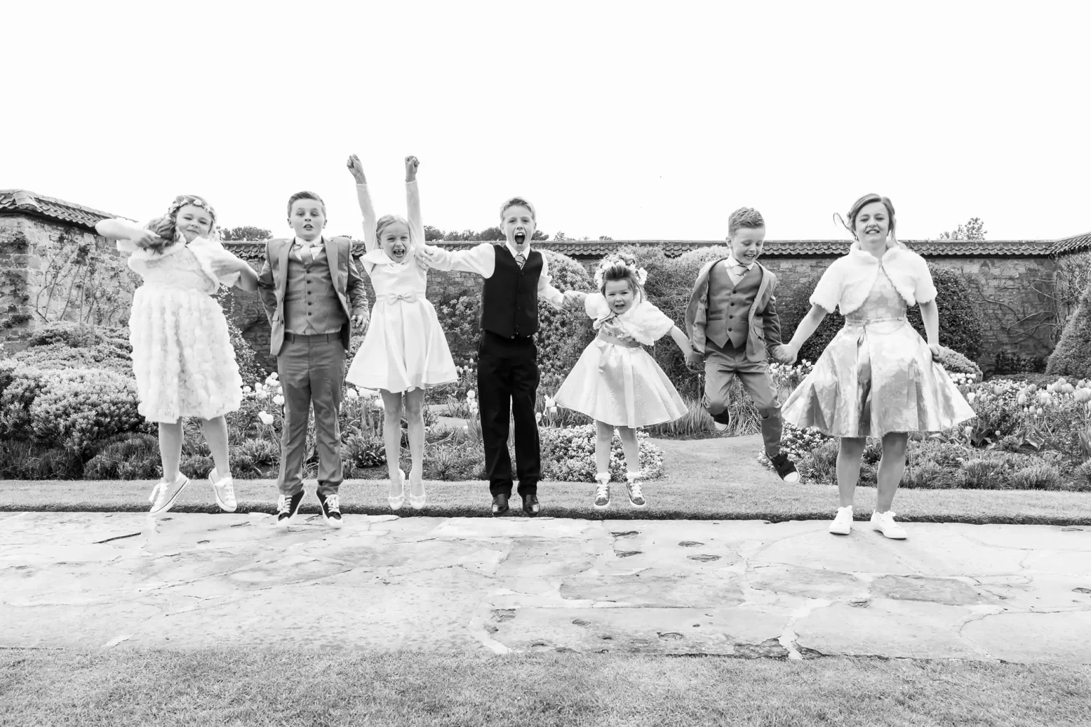 Seven children in formal attire are jumping in the air, outside on a stone path with a garden backdrop.