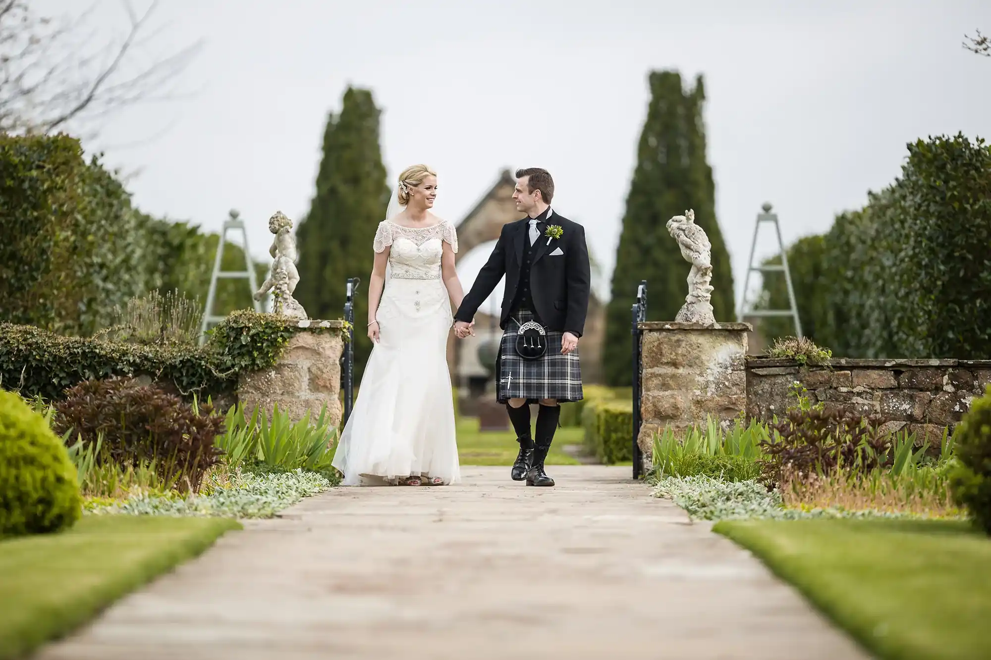 A bride in a white dress and a groom in a kilt holding hands while walking down a garden path, with statues on pedestals on both sides.