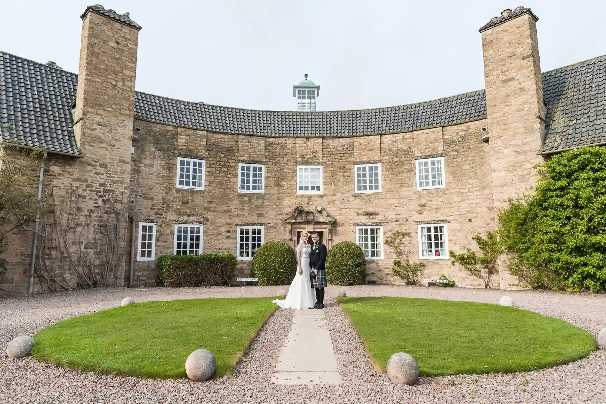A bride and groom holding hands in front of a large, historic stone building with a green lawn and symmetrical gravel pathway.