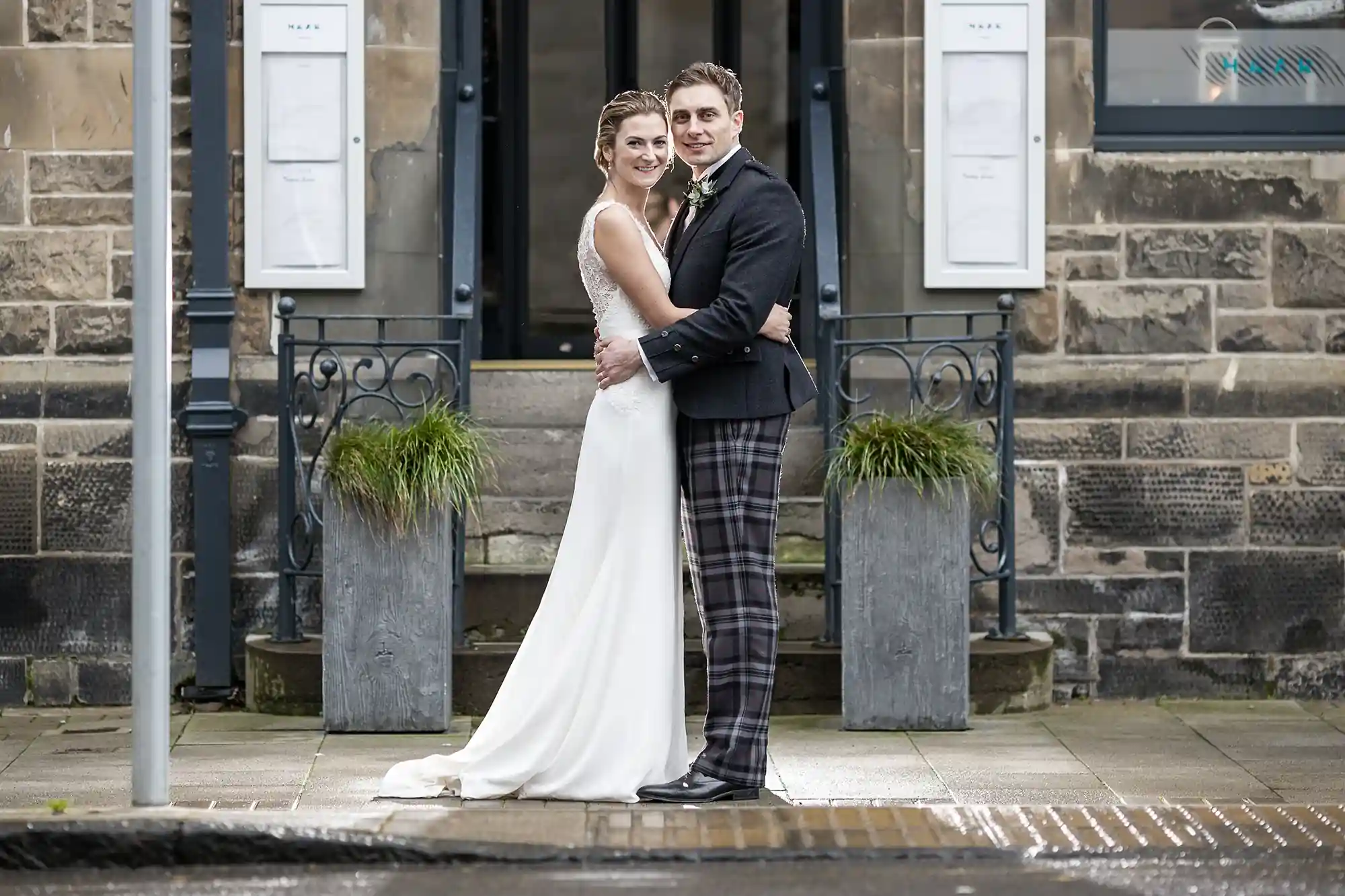 A bride in a white dress and a groom in a kilt embracing outside a building with classical architecture.