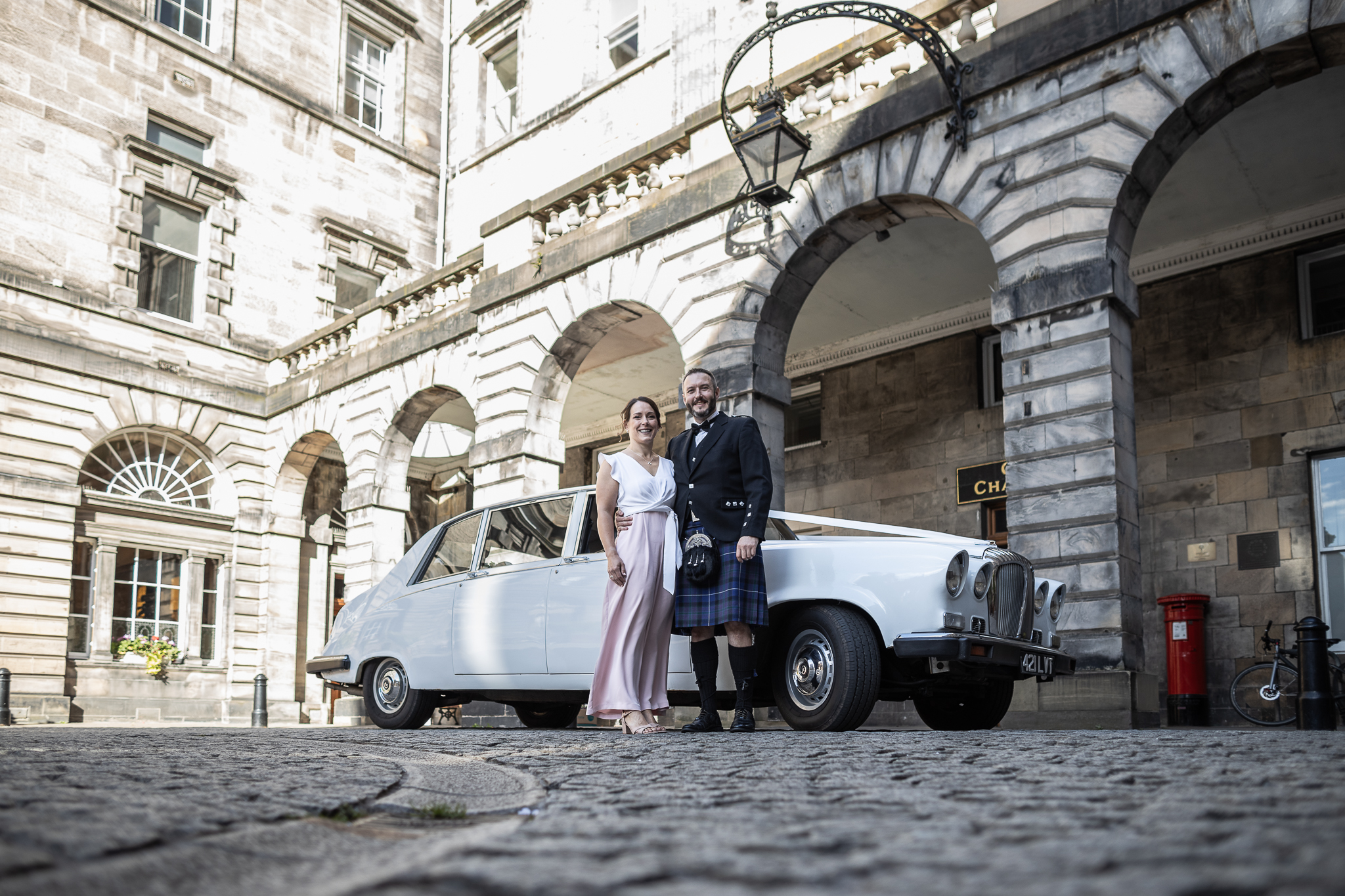 A couple poses in front of a vintage white car, with an ornate building featuring arched doorways in the background. The man is wearing a kilt, and the woman is in a pink and white outfit.