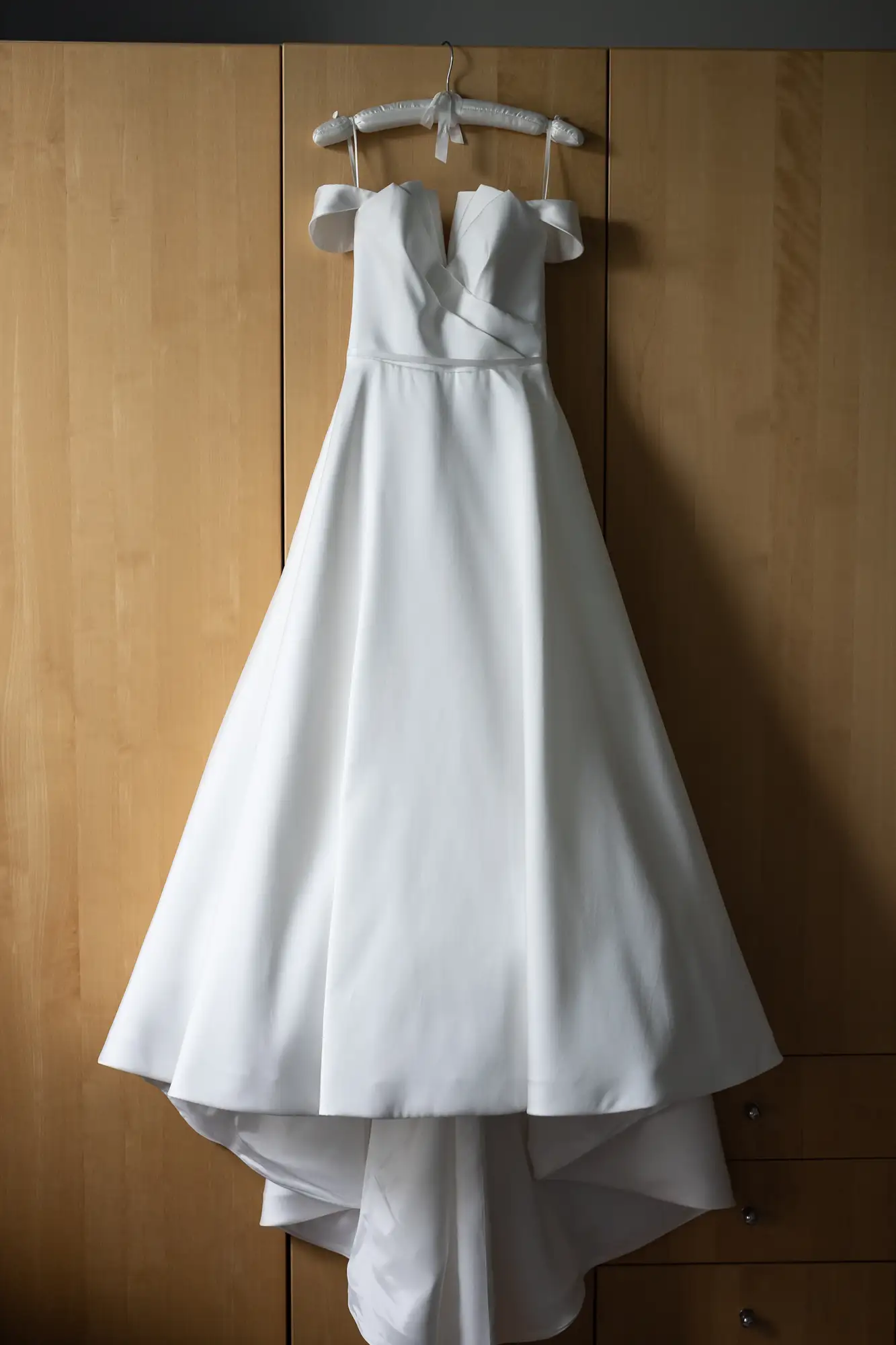 A white wedding dress with a full skirt and tailored bodice hangs on a wooden hanger against a wooden closet door.