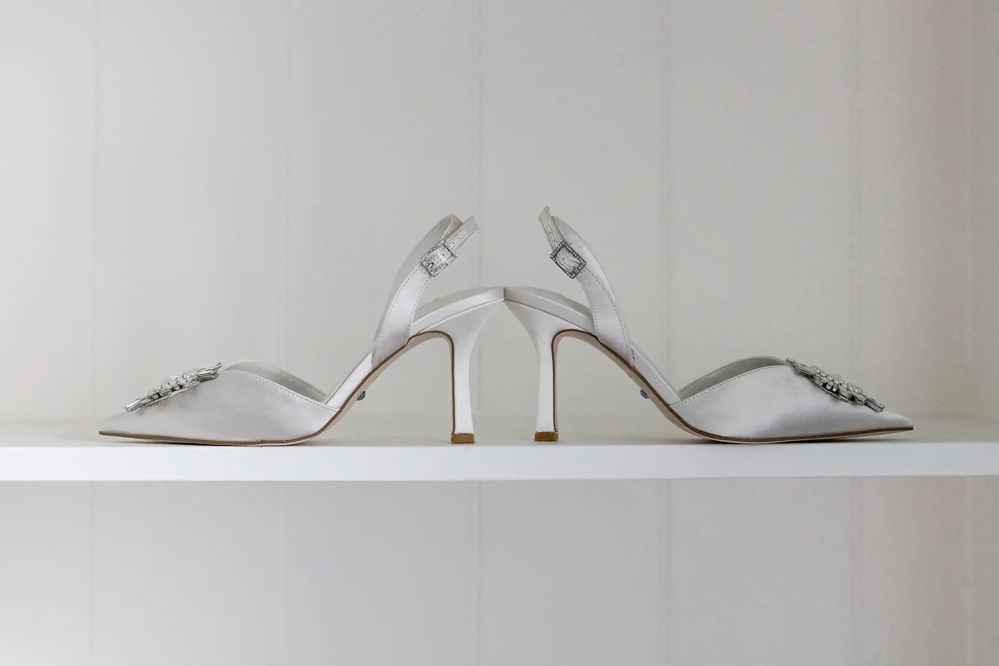 A pair of elegant white high-heeled sandals with sparkling gemstone embellishments on the straps, displayed on a white shelf.
