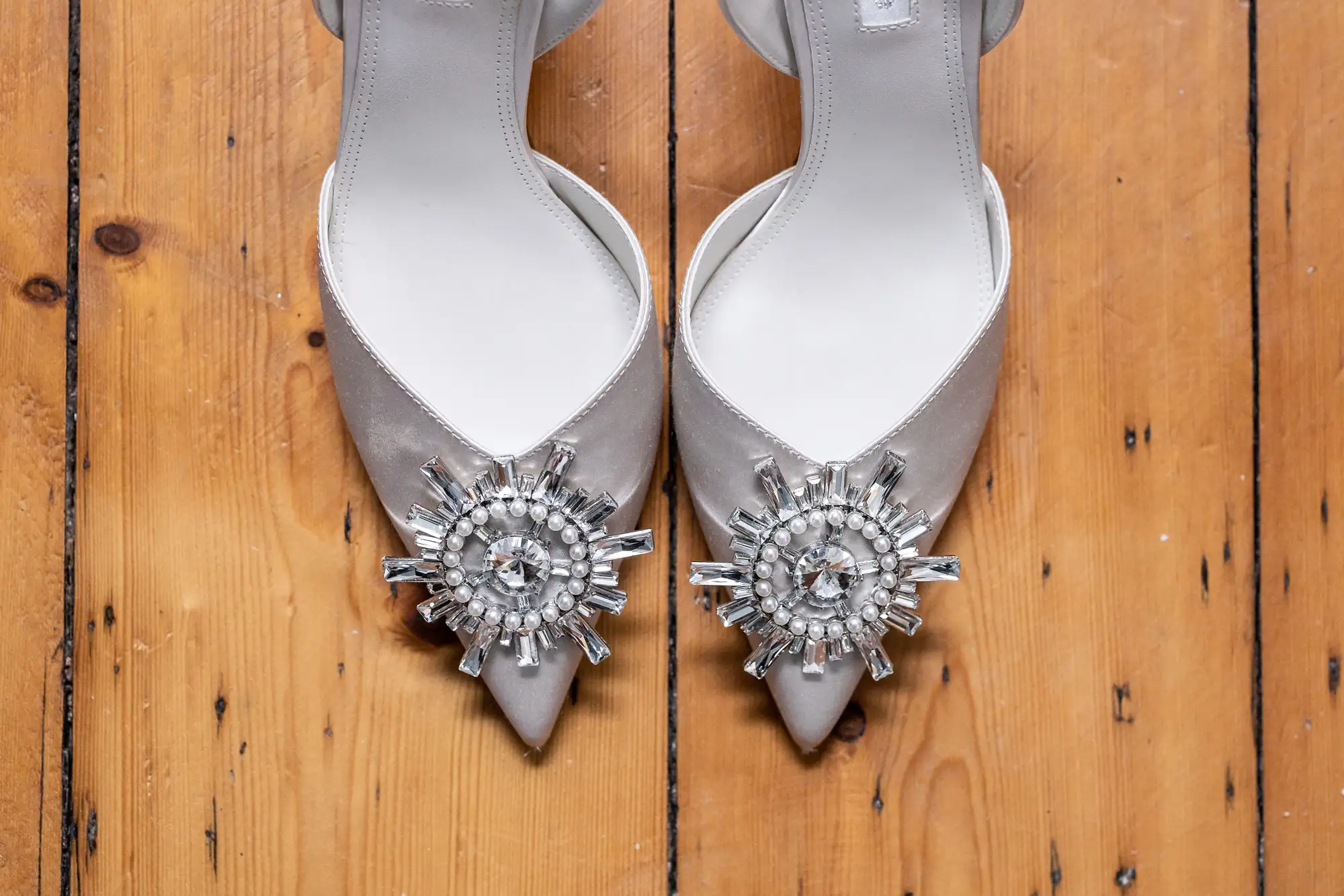 A pair of silver high-heeled shoes with decorative rhinestone embellishments on a wooden floor.