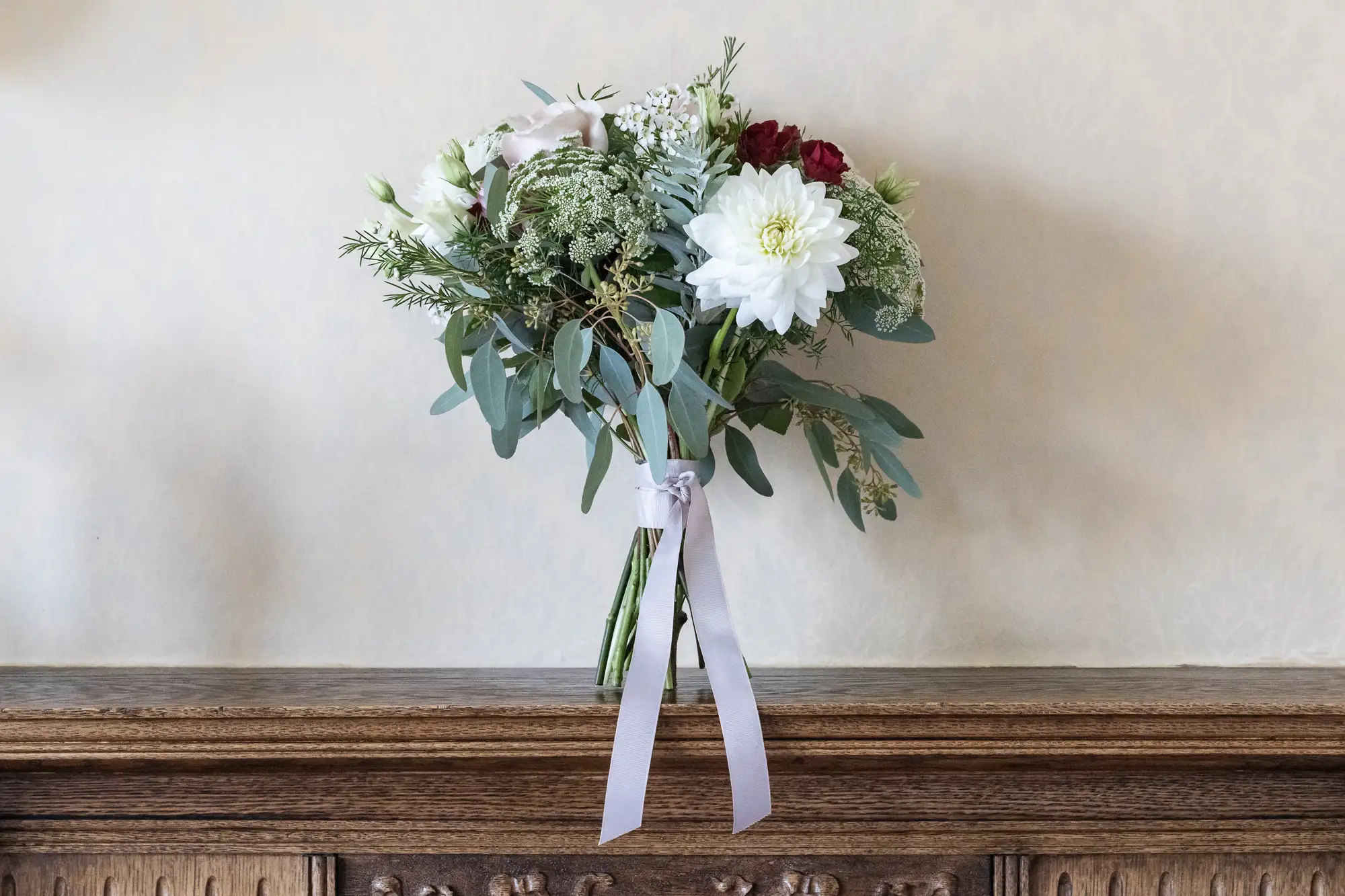 A bouquet of flowers including white, pink, and red blooms with green foliage, tied with a pink ribbon, resting on a wooden ledge against a cream wall.
