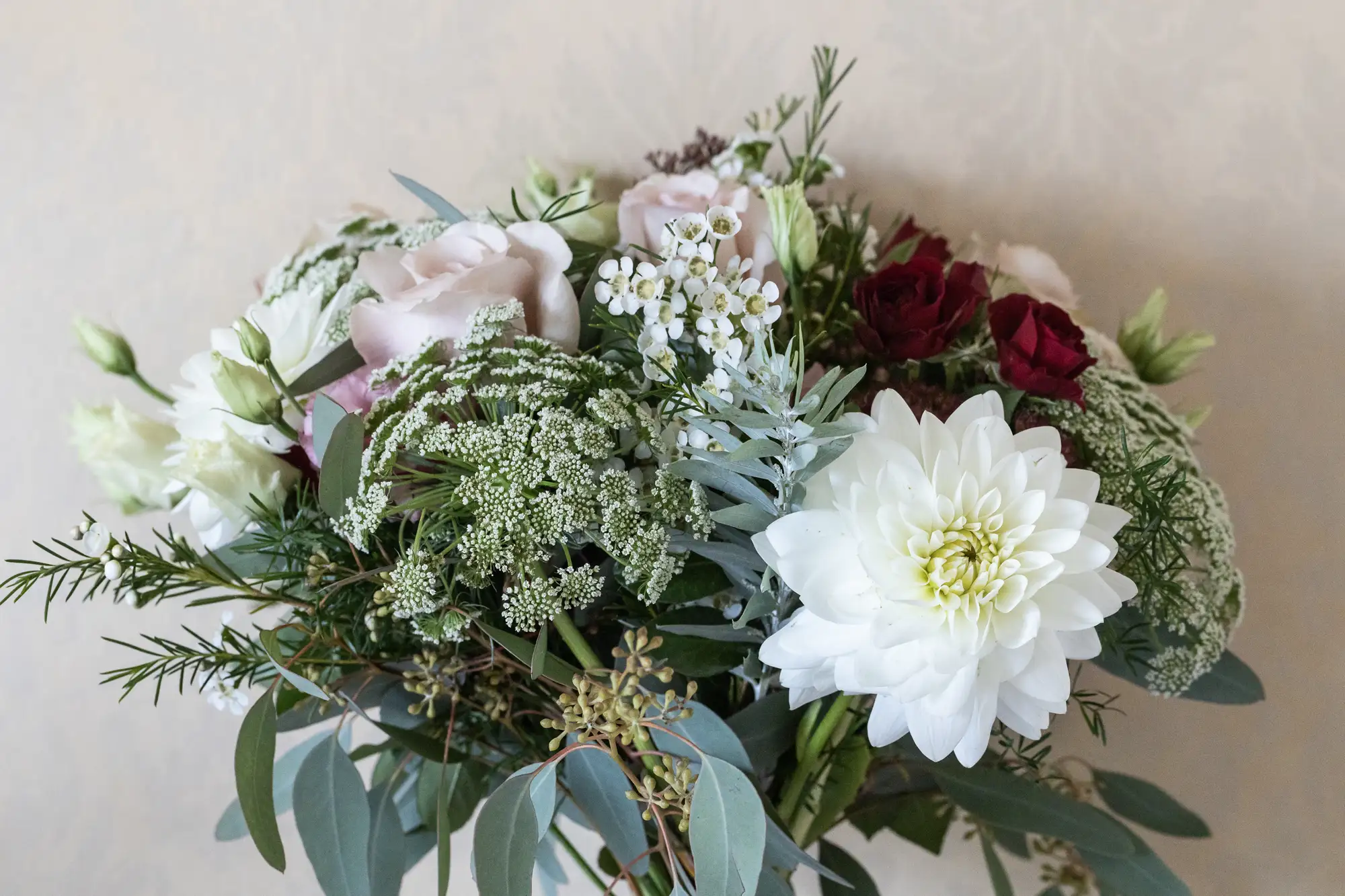 A floral arrangement featuring white dahlias, deep red roses, pink blooms, baby's breath, and various greenery on a light textured background.