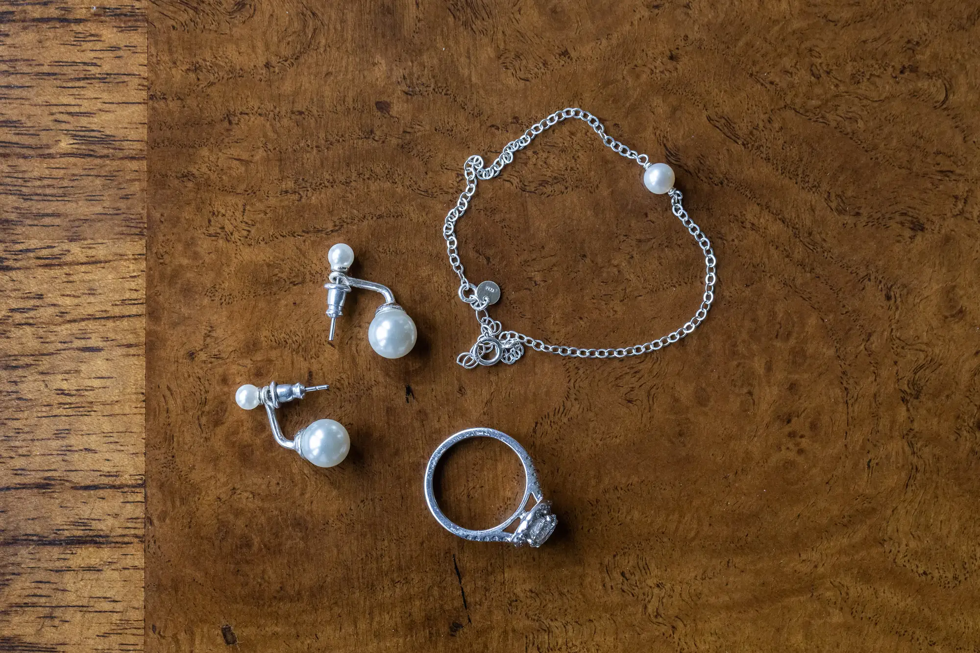 Pearl earrings, a silver bracelet with a heart charm, and a diamond ring displayed on a wooden surface.