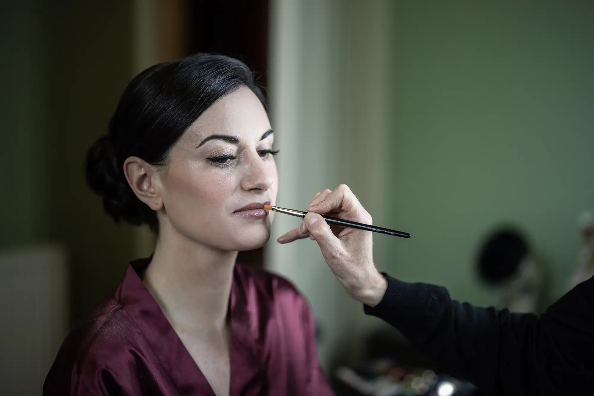 A woman in a satin robe having makeup applied by a makeup artist using a brush, in a softly lit room.