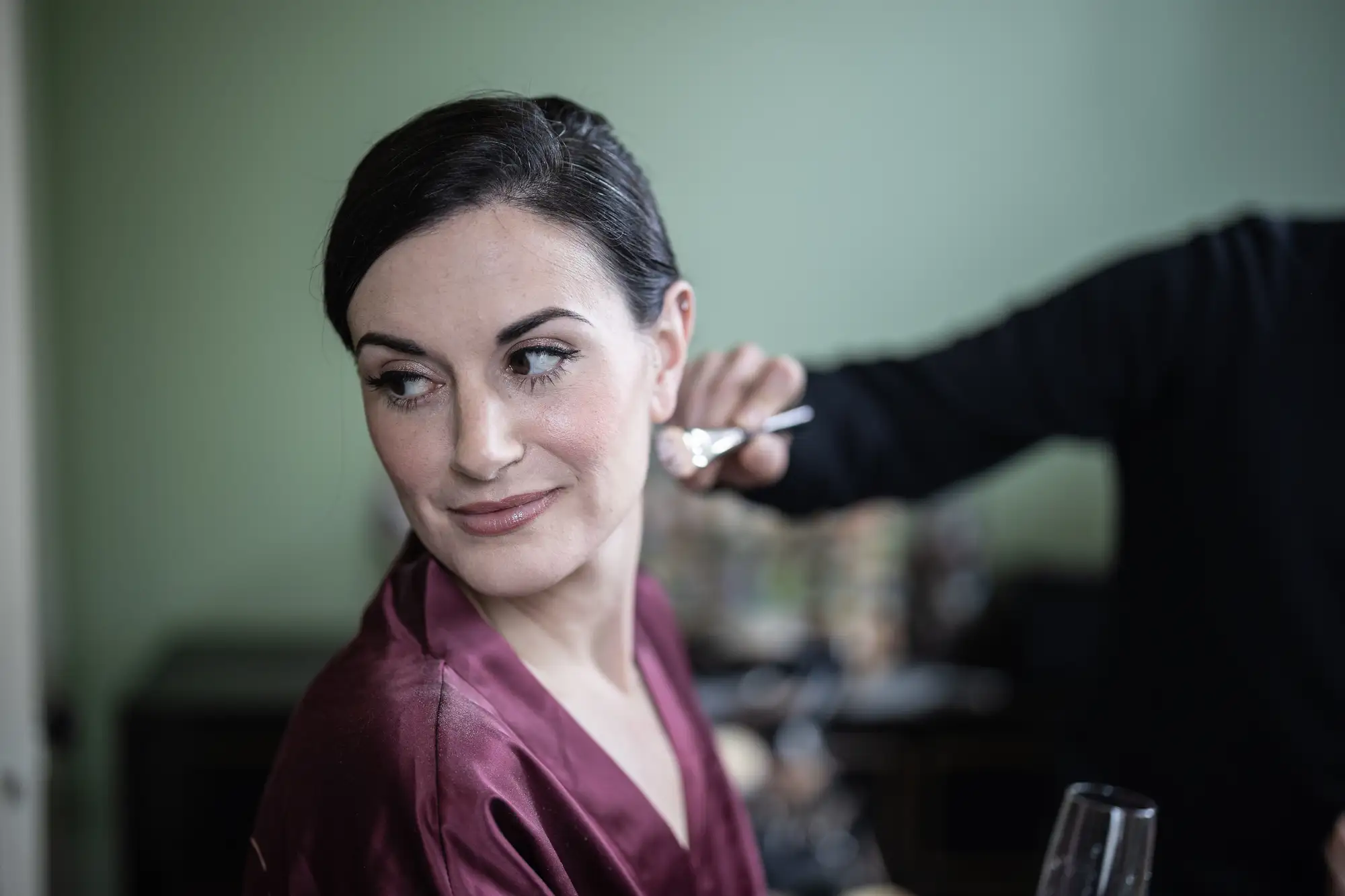 A woman in a maroon robe smiles as a makeup artist applies makeup to her face in a room with a green wall.