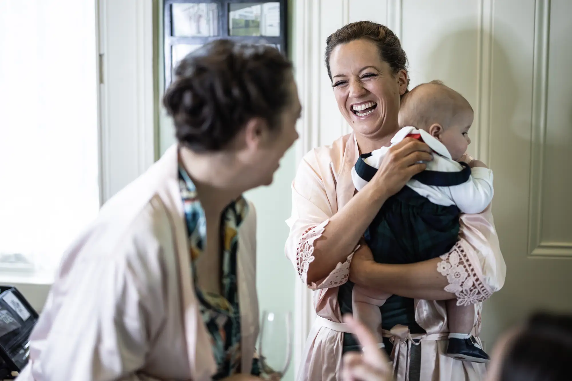 A woman in a pink dress smiles joyfully while holding a baby, conversing with another woman in a cafe setting.