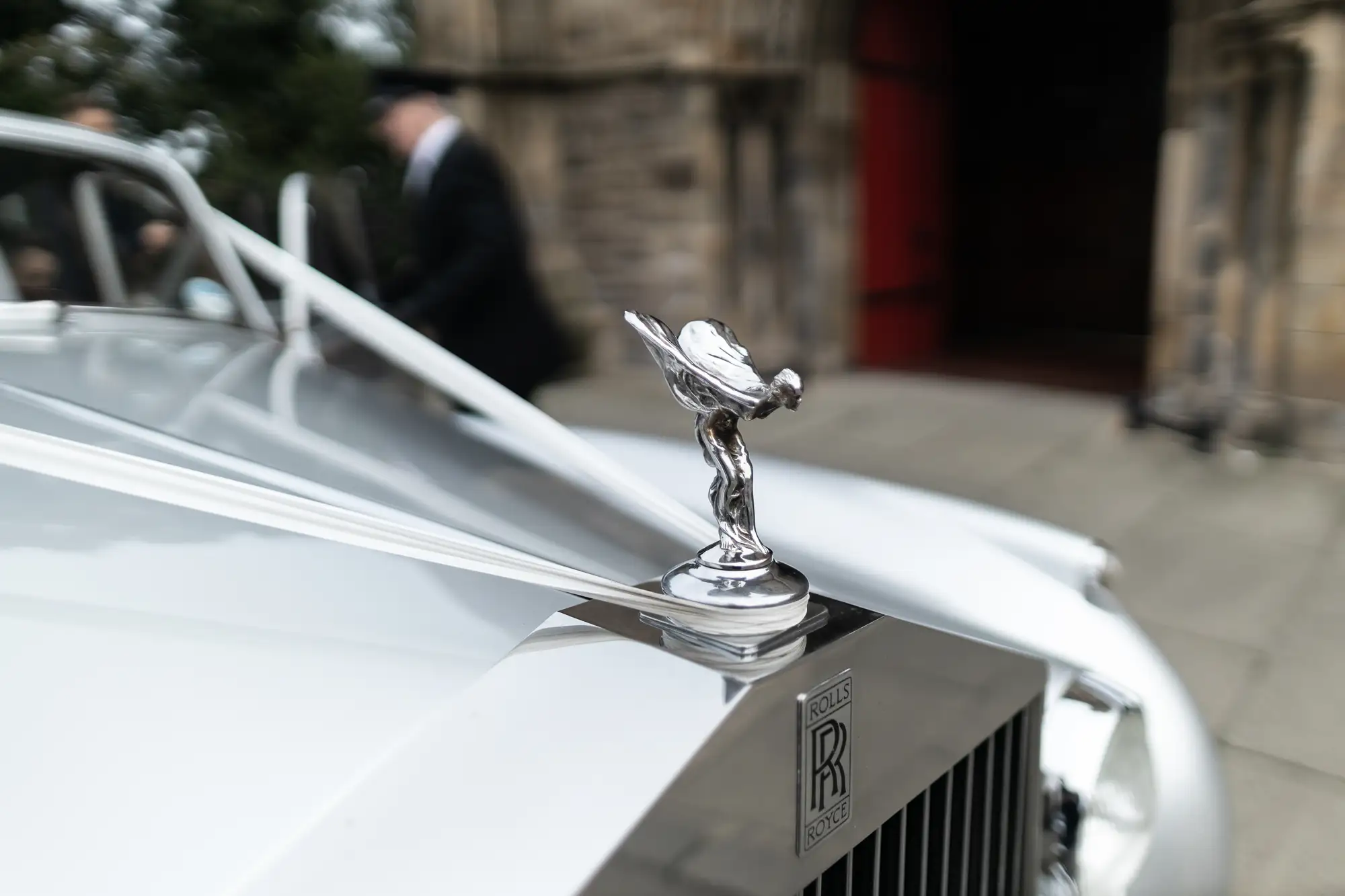 A rolls-royce hood ornament, featuring the iconic "spirit of ecstasy," mounted on the front of a white car with a blurry figure in the background.