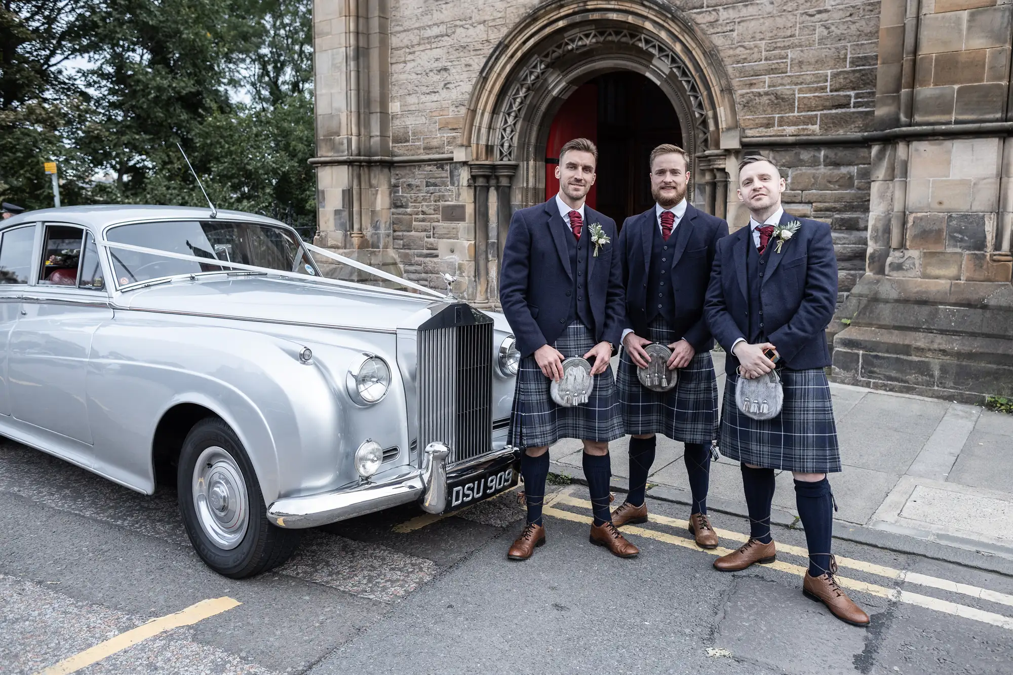 Three men in kilts and blazers stand by a classic silver rolls-royce near a stone church, each holding a glass.