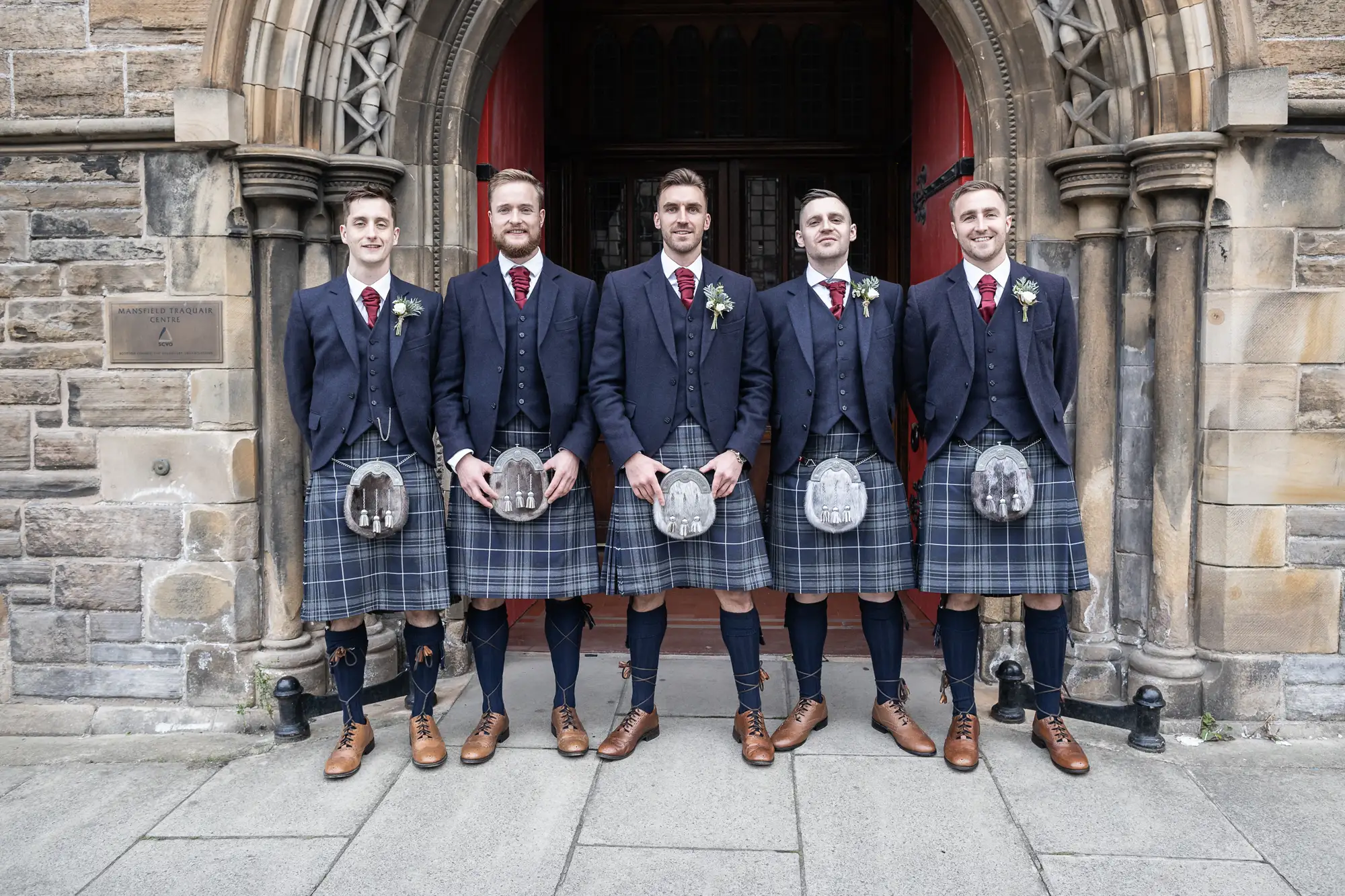 Five men in traditional scottish kilt attire standing in front of a church entrance, each holding a sporran and wearing a boutonniere.