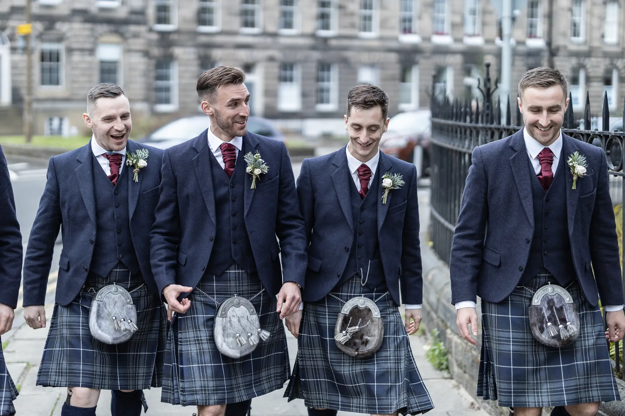 Four men in tartan kilts and jackets, laughing and walking together, each wearing a boutonniere and holding a sporran.