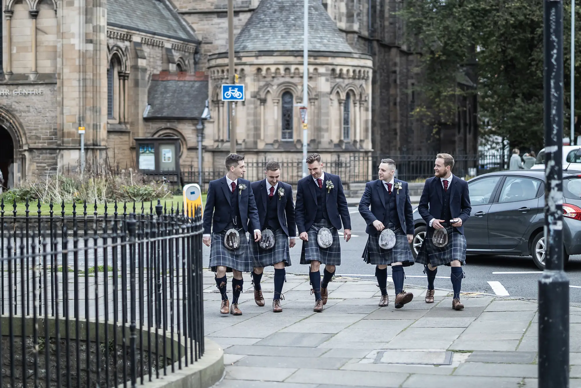 Four men in kilts and blazers walking along a city street, with historic architecture in the background.