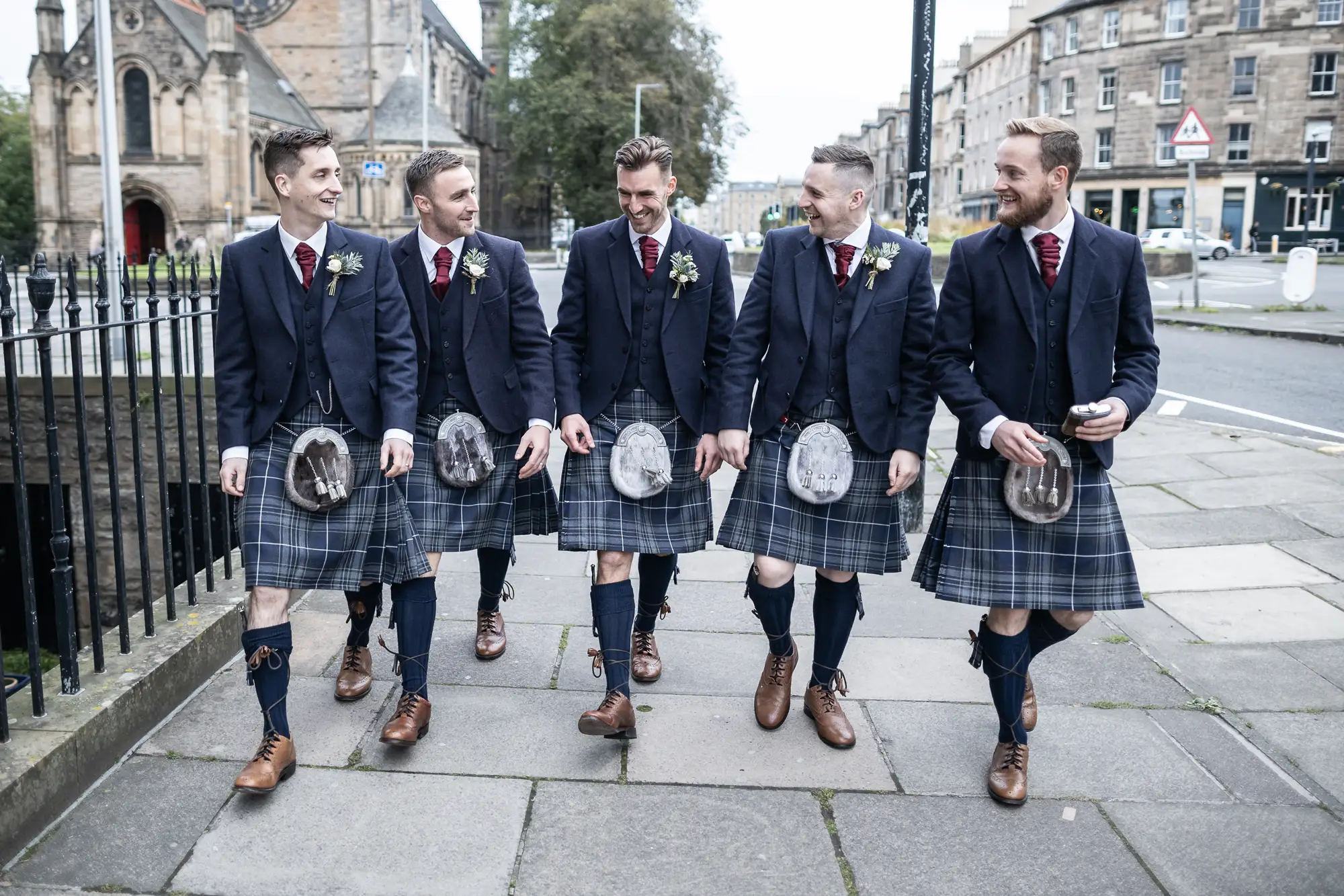 Five men in traditional scottish attire with kilts and sporrans walking and laughing together on a city street.