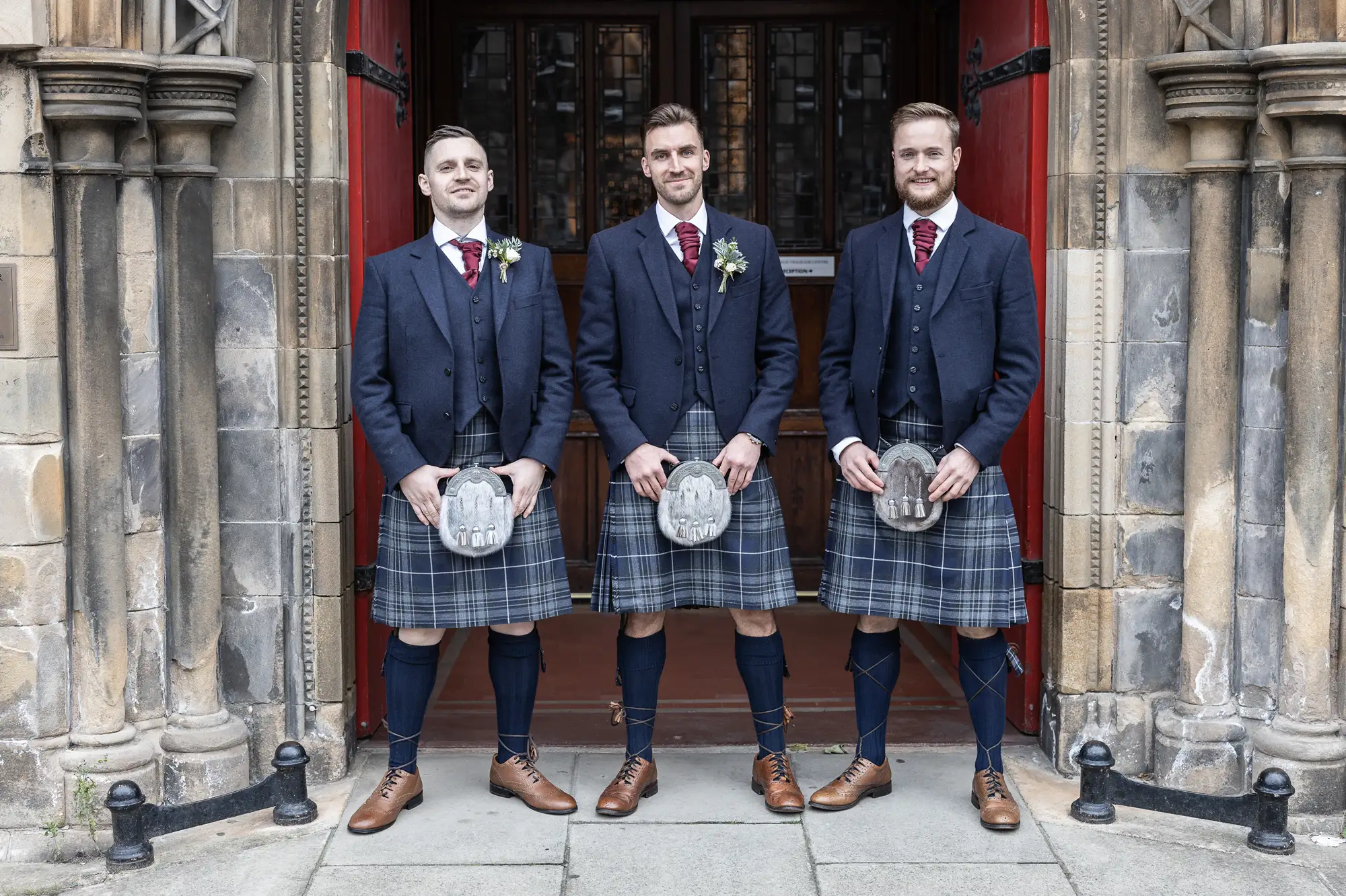 Three men in traditional scottish kilts and jackets stand in front of a church's ornate red doors, each wearing a boutonniere and holding a sporran.