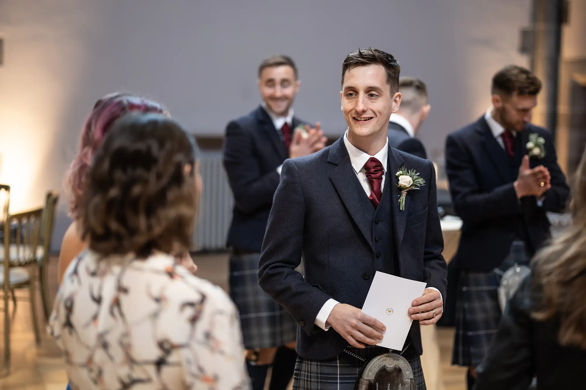 A groom in a kilt smiles at a bride, holding a document, with applauding guests in the background at a wedding ceremony.