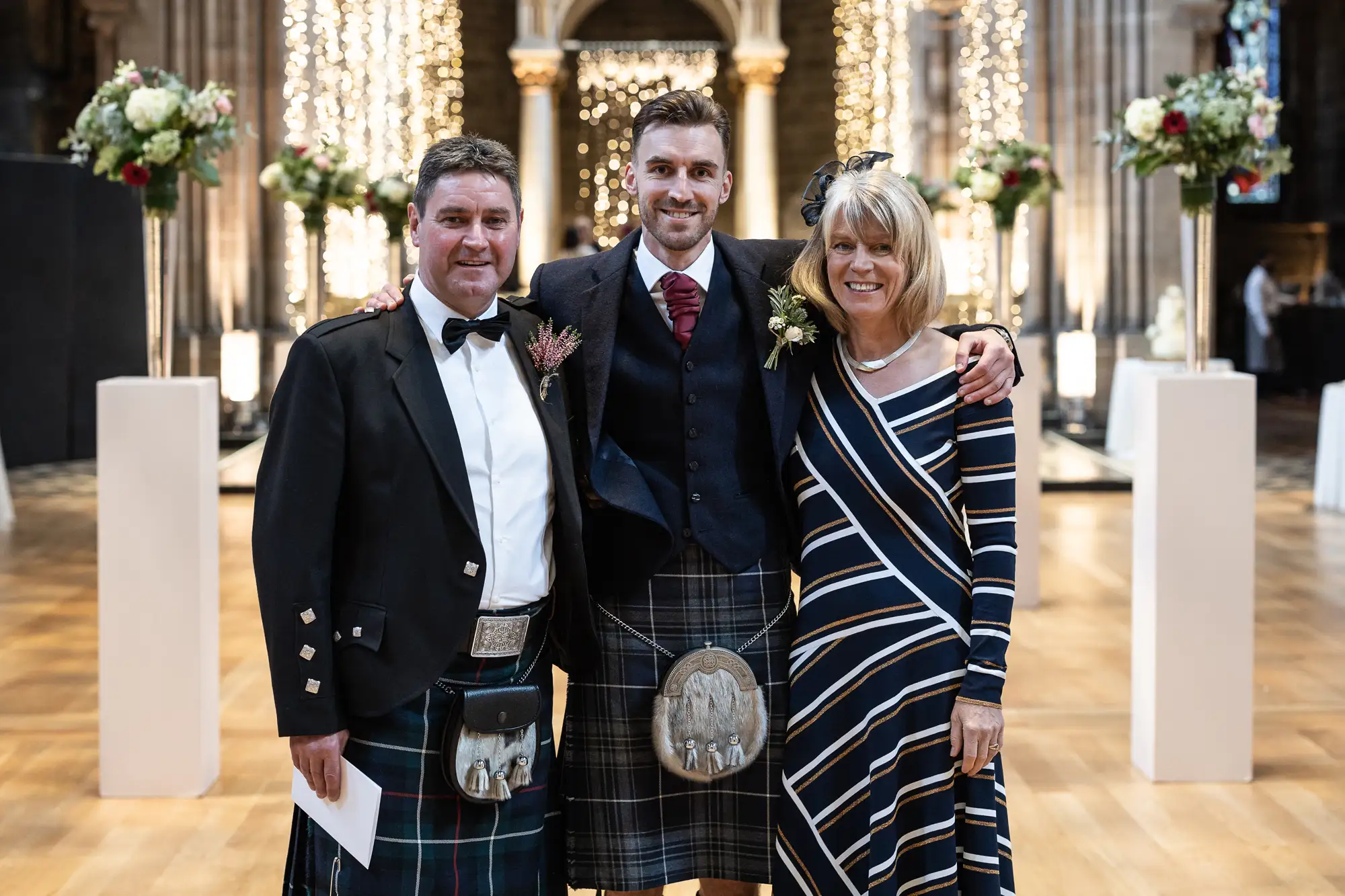 Three people in formal attire pose in a cathedral; two men in kilts flank a woman in a striped dress, all smiling.