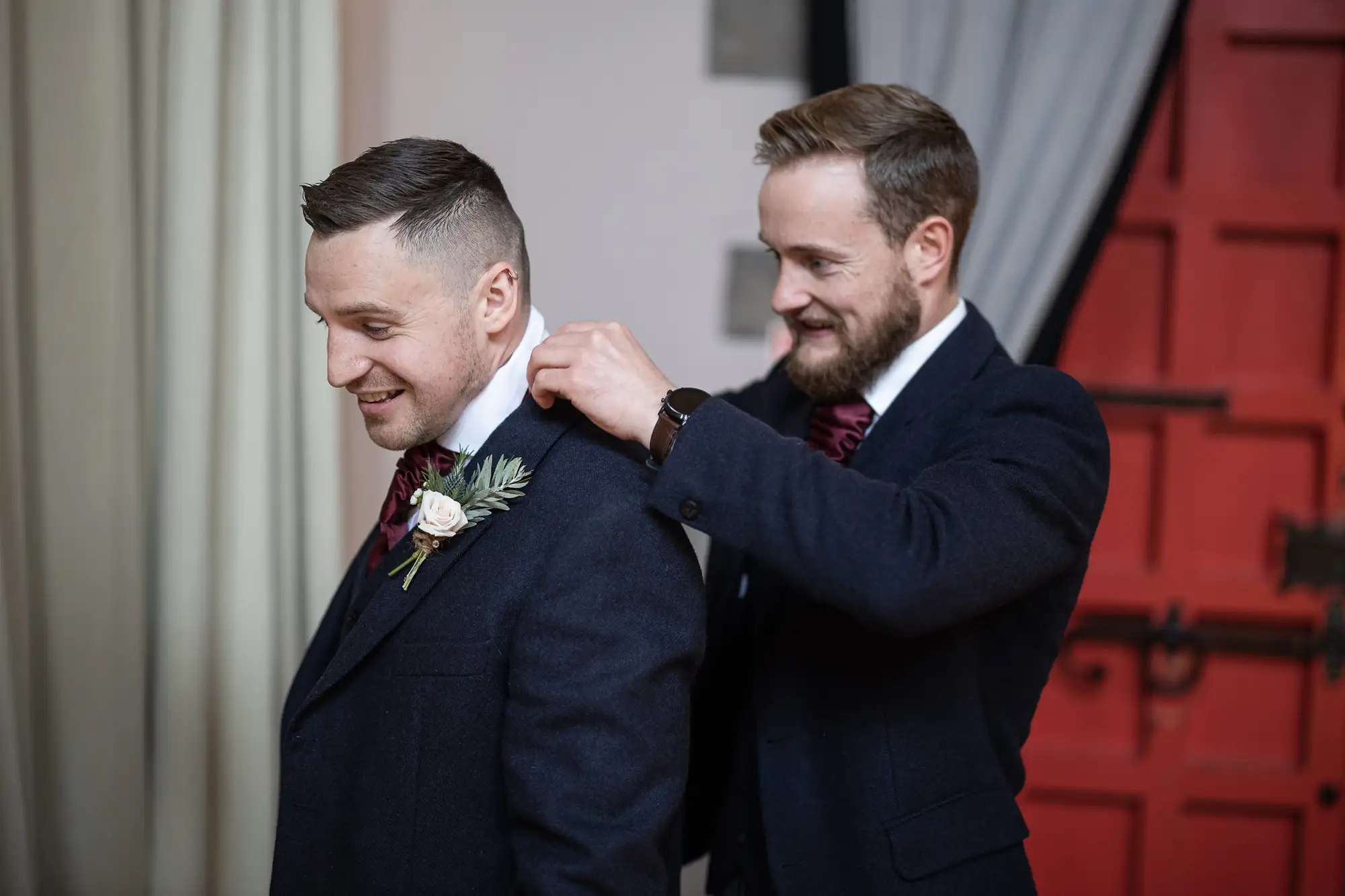 A man adjusts the boutonniere on the lapel of another man's dark suit in an elegant room, both smiling subtly.