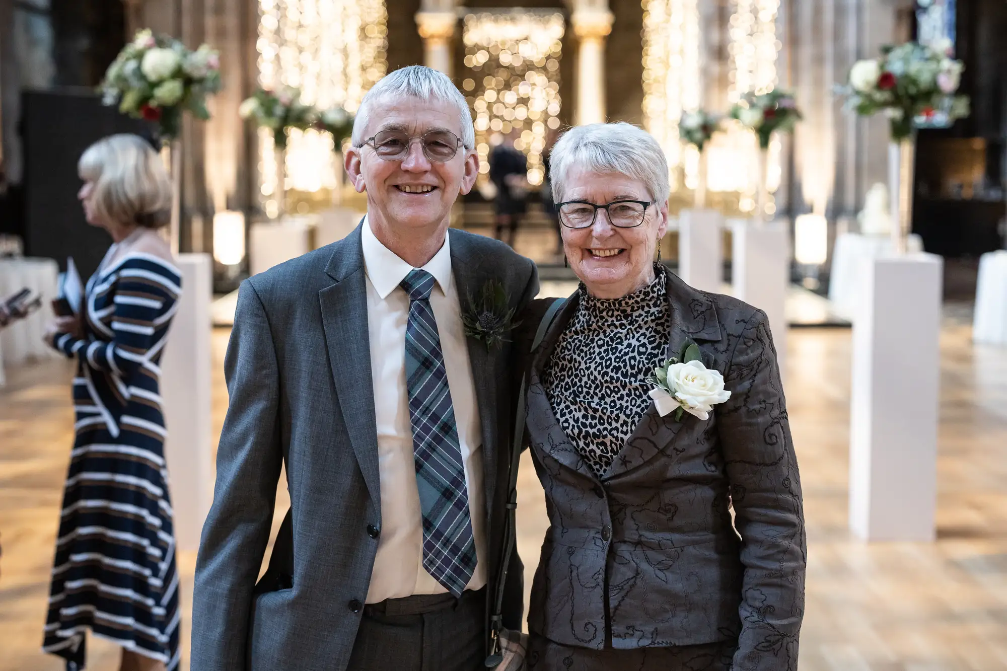 An elderly couple smiling at a wedding, the man in a gray suit with a boutonniere and the woman in a patterned dress with a corsage, inside a grand hall with floral decorations.